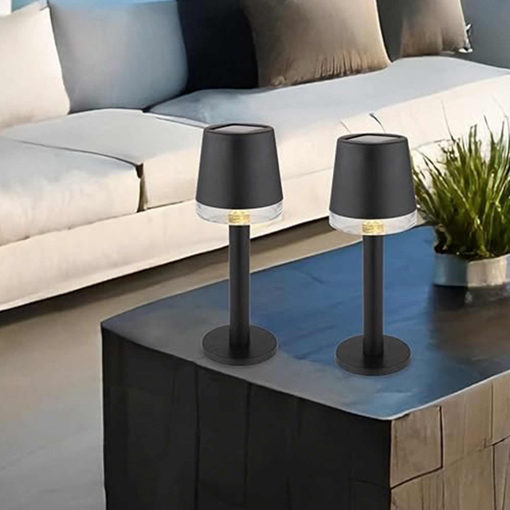 Dos lámparas negras con bases transparentes sobre una mesa oscura. Un sofá está en el fondo con almohadas y una planta.