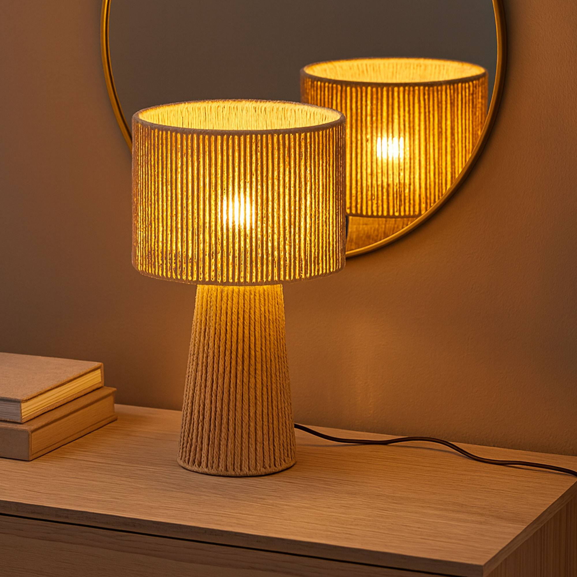 Una lámpara encendida con pantalla y base tejidas está sobre una superficie de madera. Un espejo refleja la lámpara. Libros a la izquierda.