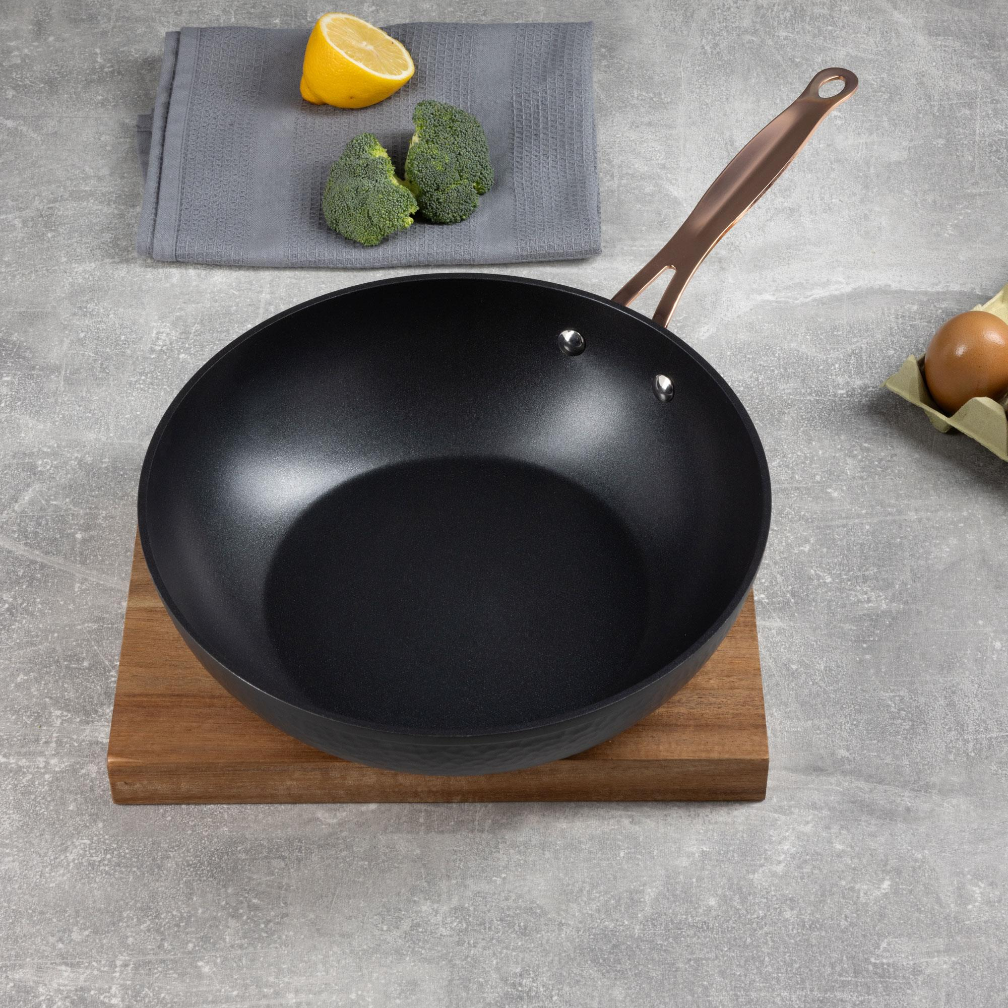 Una sartén wok sobre una tabla de madera con limón, brócoli y huevos. Mango de cobre. Fondo gris.