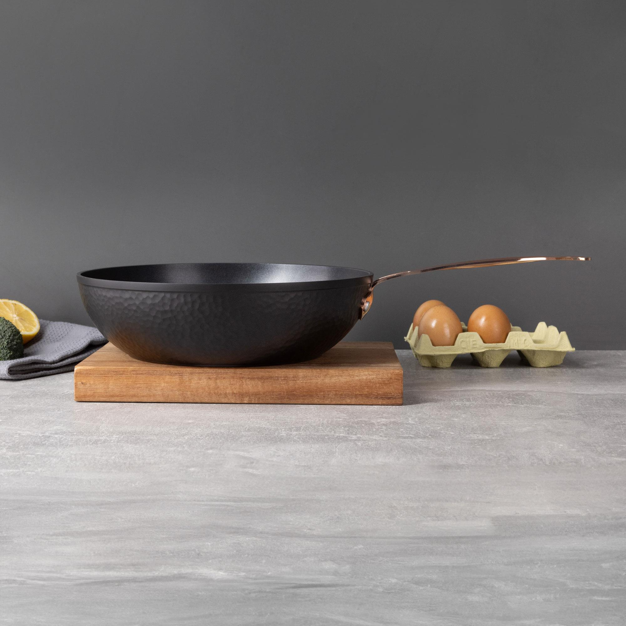 Una sartén negra sobre una tabla de madera con huevos en una caja sobre una superficie gris, con fondo gris.