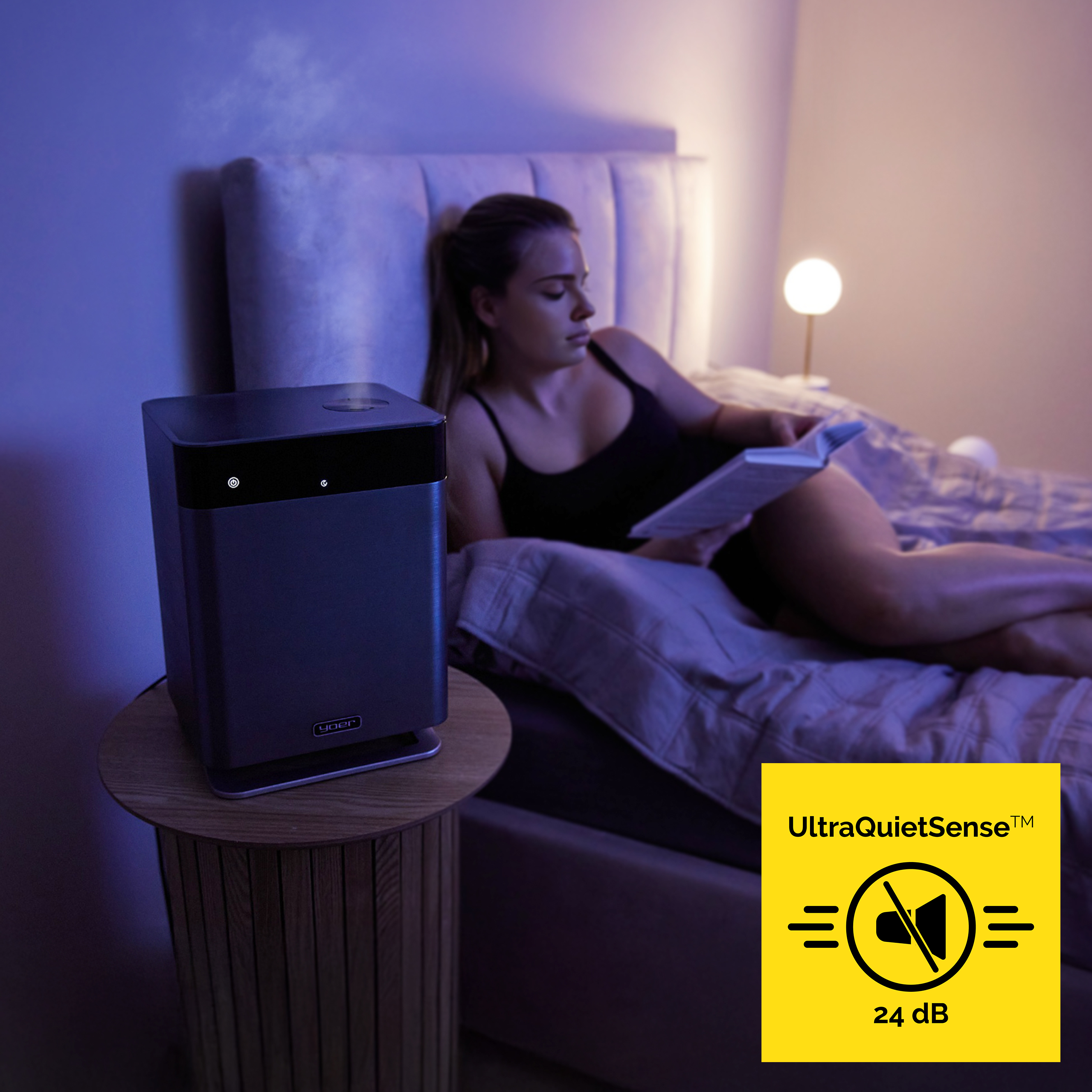 Kobieta czyta w łóżku, z oczyszczaczem powietrza na stoliku nocnym. Żółty znak wskazuje 24 dB.