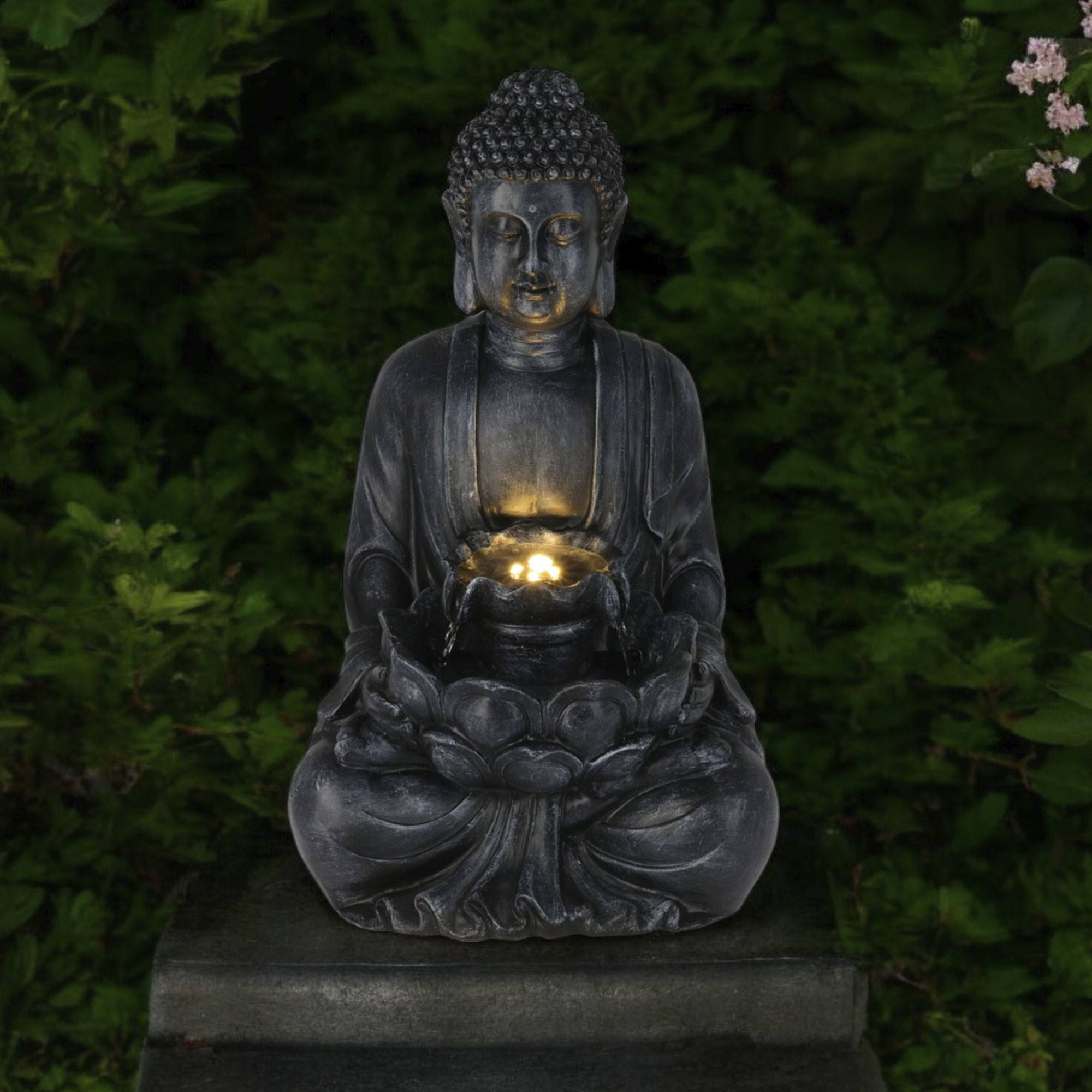 Una estatua oscura de Buda en un jardín, con agua que fluye de un cuenco central. El follaje verde forma el fondo.