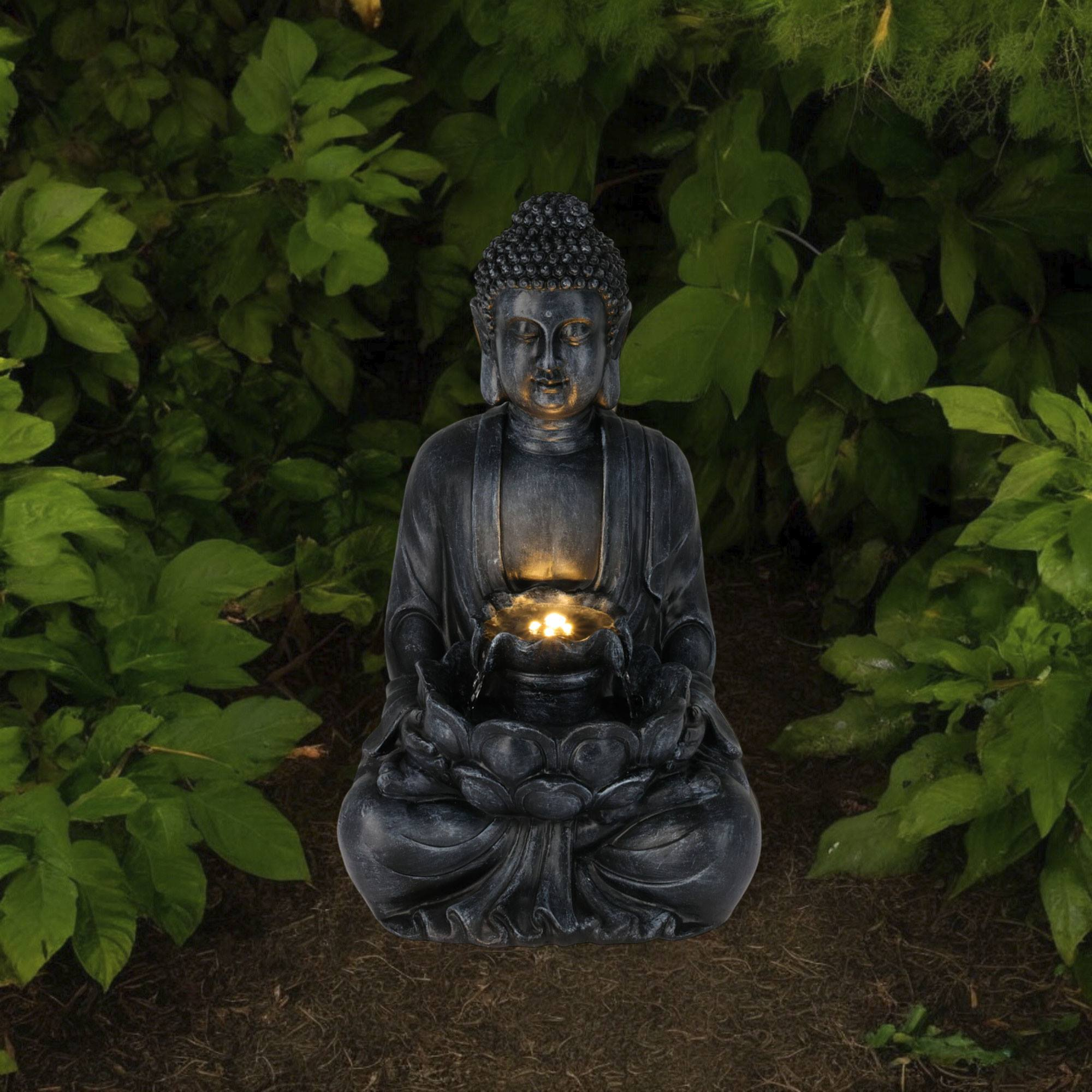 Estatua oscura de Buda en un jardín, rodeada de hojas verdes. El agua fluye del loto en las manos de la estatua.