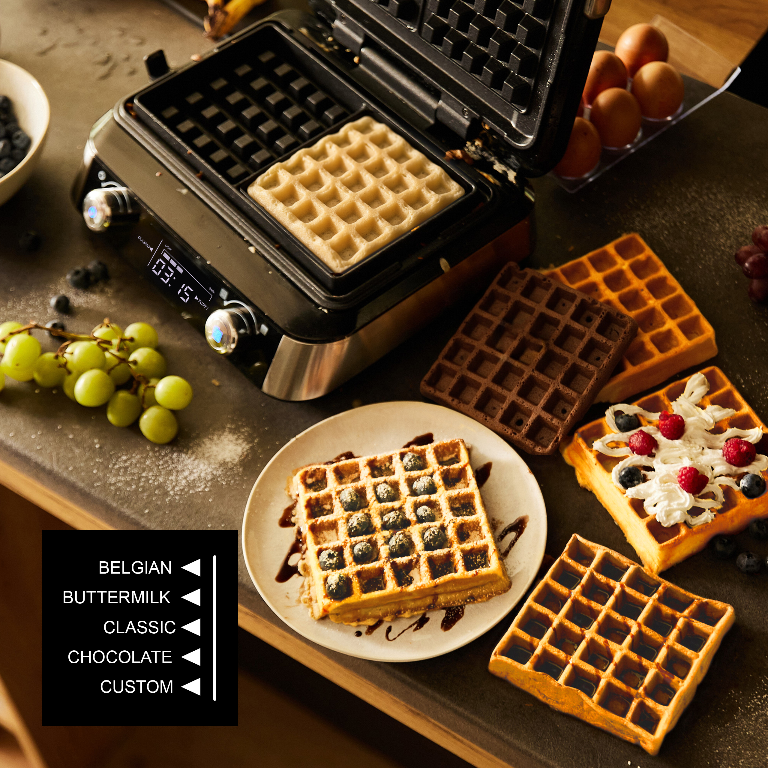 Gofrownica z gofrem w środku, otoczona upieczonymi goframi, winogronami, borówkami i jajkami.