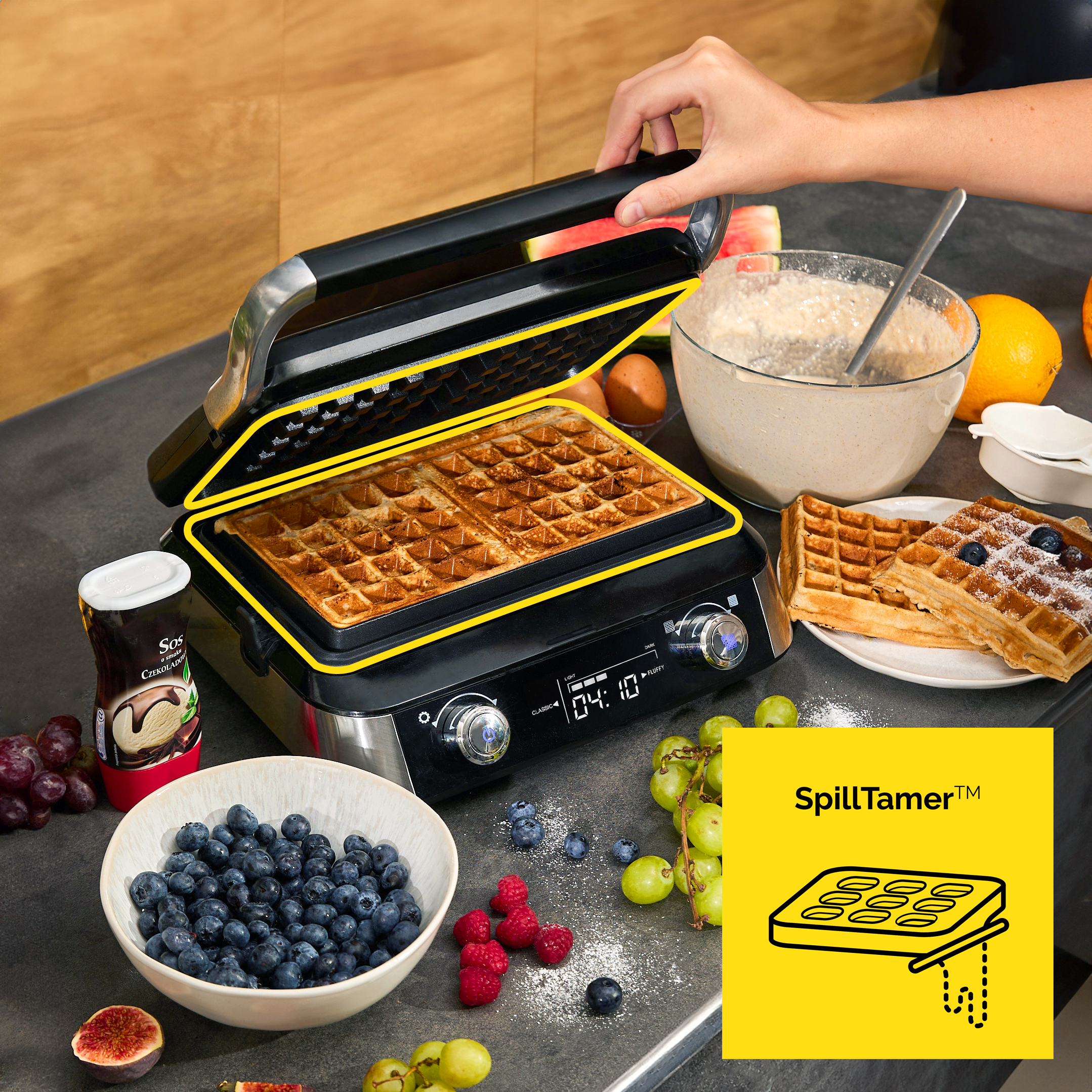 Gofrownica z upieczonymi goframi, miską z ciastem, owocami i ręką otwierającą pokrywę. Składniki gotowe do serwowania.