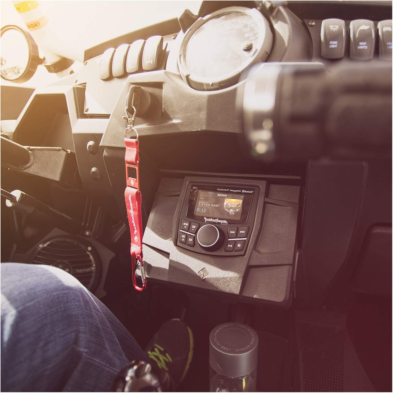 Innenansicht des Autos. Armaturenbrett mit Radio, Anzeigen, Schaltern und rotem Schlüsselband. Bein und Schuh sichtbar. Wasserflasche im Vordergrund.