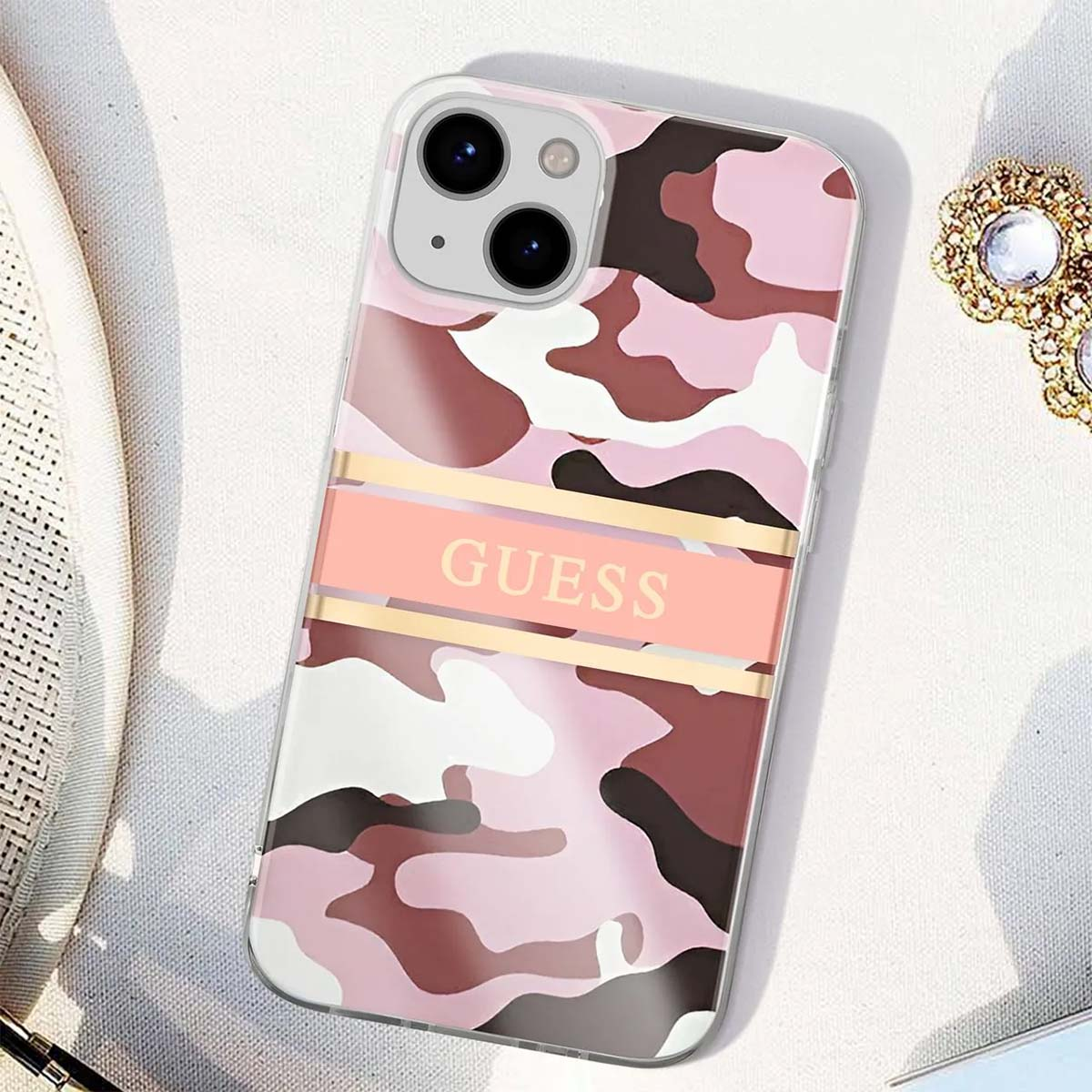 Custodia per telefono rosa camouflage con la scritta GUESS. Si trova su una superficie bianca con gioielli sullo sfondo.