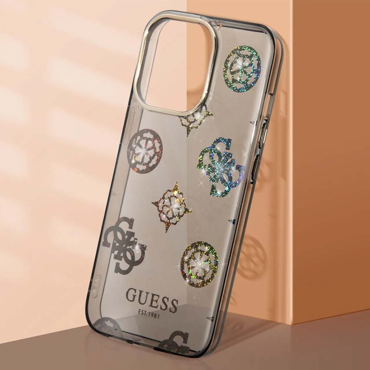 Una custodia trasparente Guess con disegni glitterati e logo. Poggia su una superficie con sfondo color pesca.