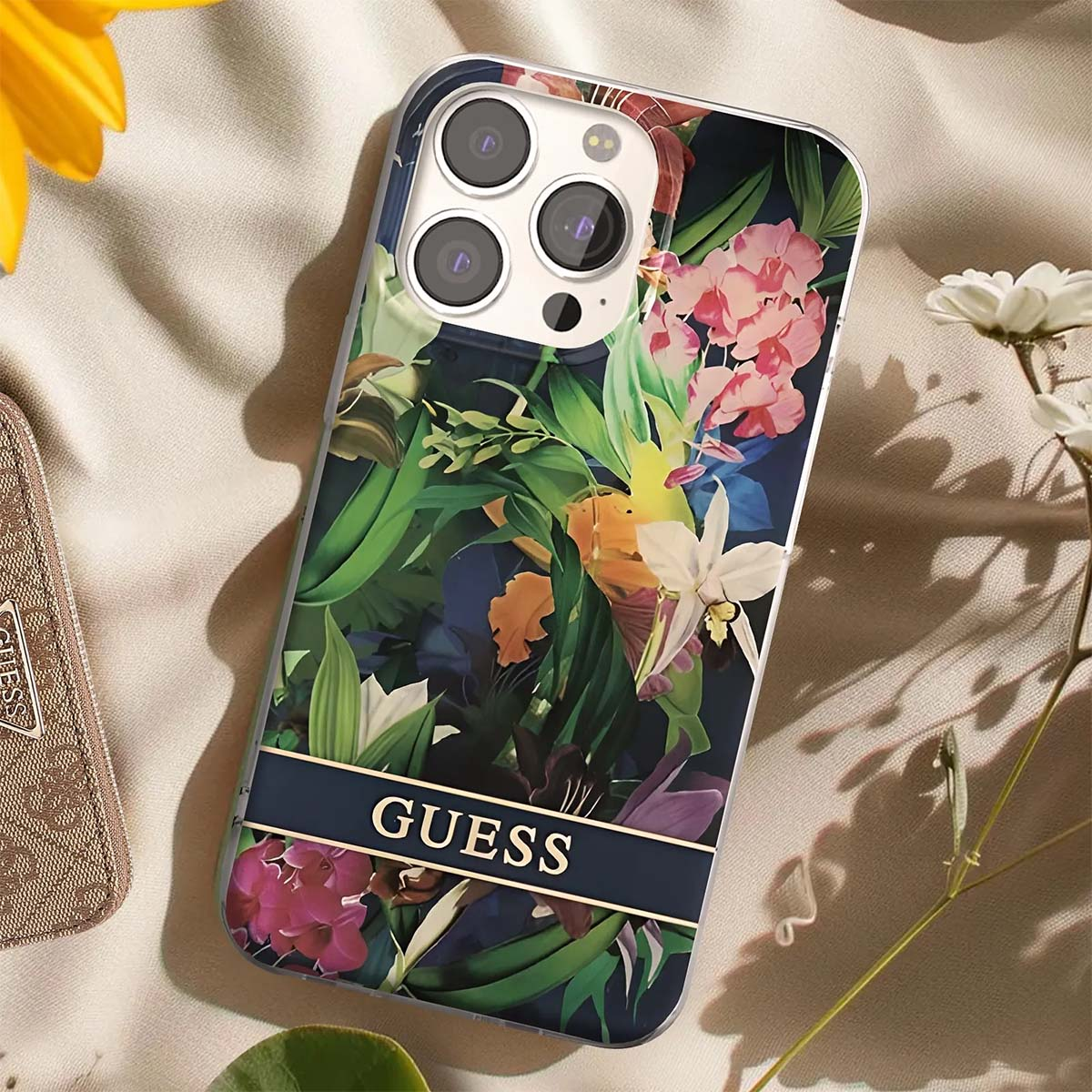 Una custodia per telefono con un motivo floreale, con fiori rosa, bianchi e verdi e la scritta 'GUESS'. Lo sfondo è beige.
