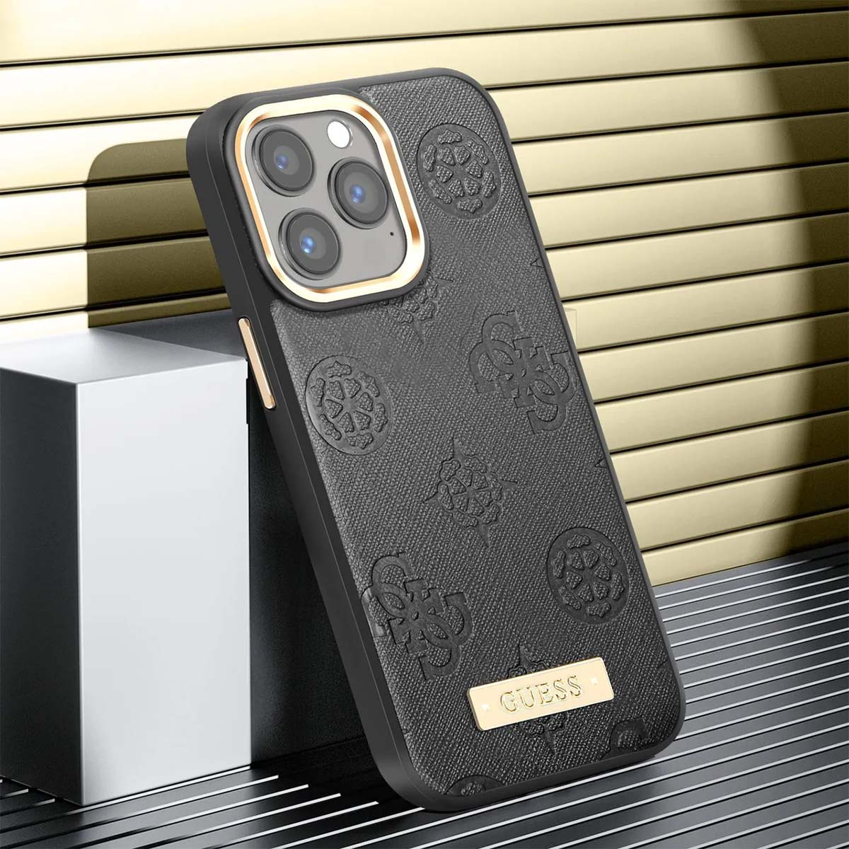 Una custodia per telefono Guess nera con motivi in rilievo. Accenti dorati incorniciano la fotocamera e il logo. Lo sfondo è dorato.