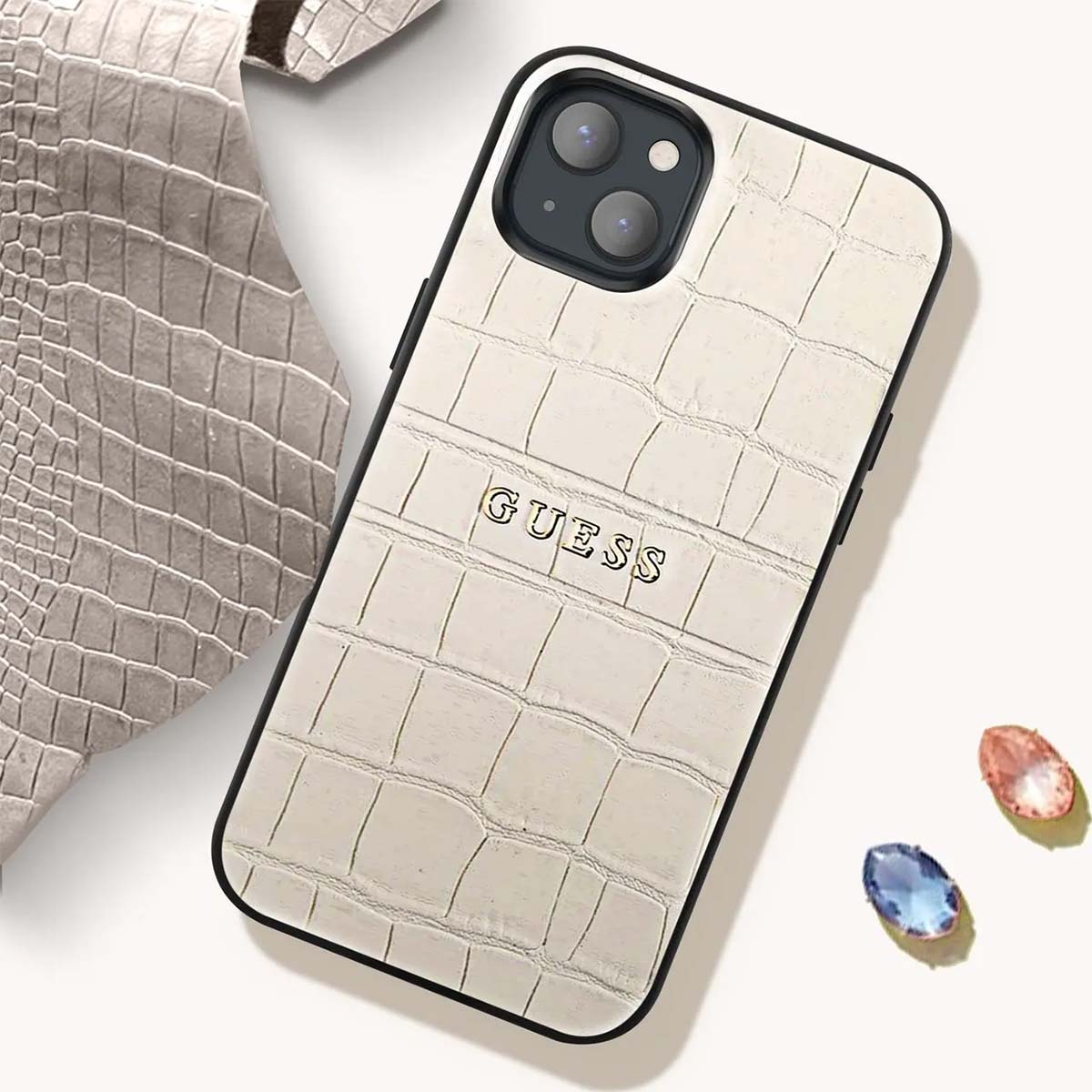 Una custodia per telefono Guess bianca con motivo a coccodrillo. Un pezzo di pelle grigia è accanto. Sono visibili due gemme.