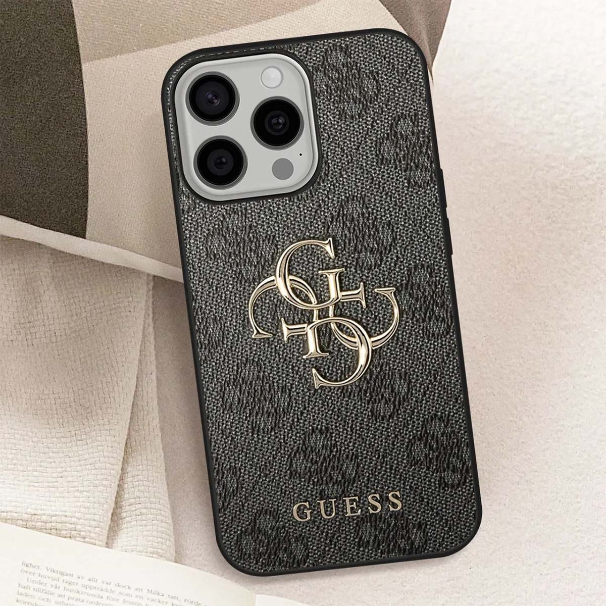 Eine dunkelgraue Guess-Handyhülle mit goldenem Logo und Branding. Die Handyhülle liegt auf einer hellbeigen Oberfläche.