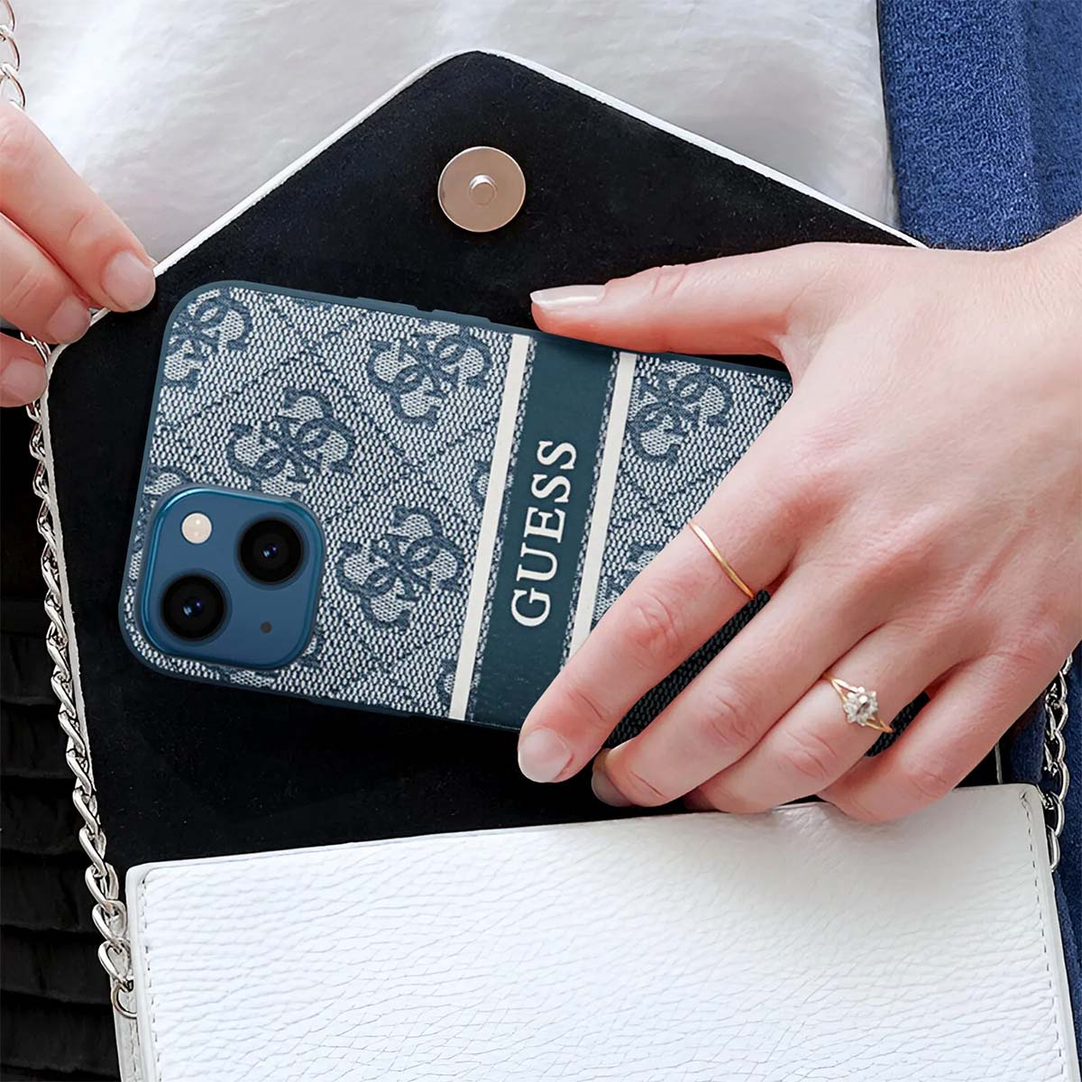 Eine Person hält ein Telefon in einer Guess-Hülle über einer weißen Handtasche. Das Gehäuse ist blau mit einem Muster und dem Wort Guess.