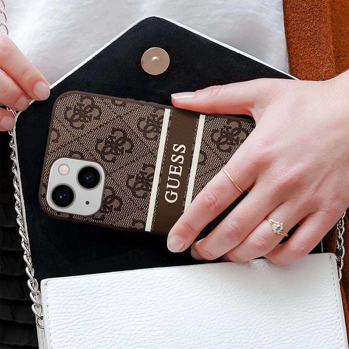 Una donna che tiene una custodia per telefono Guess in una borsa bianca. La custodia è marrone con il logo Guess. Indossa anelli.