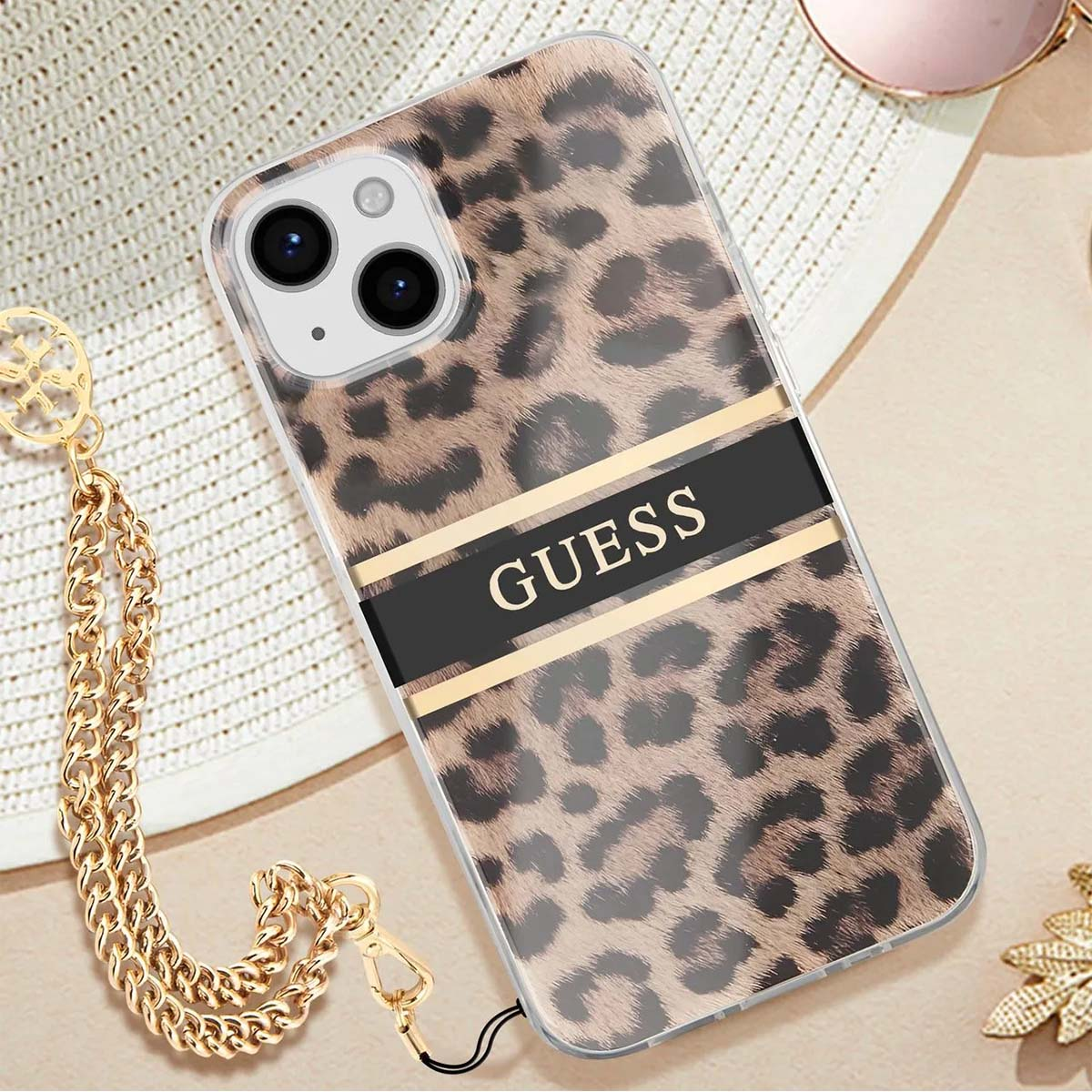 Una custodia per telefono con stampa leopardata con 'GUESS', una catena dorata e un cappello bianco sullo sfondo.