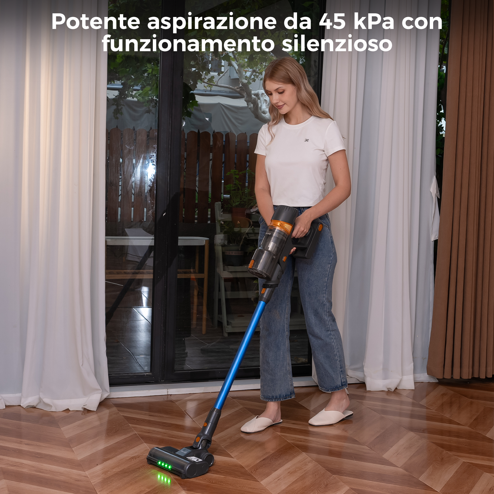 Una donna aspira un pavimento in legno con un aspirapolvere blu. Tende bianche e finestra sullo sfondo.