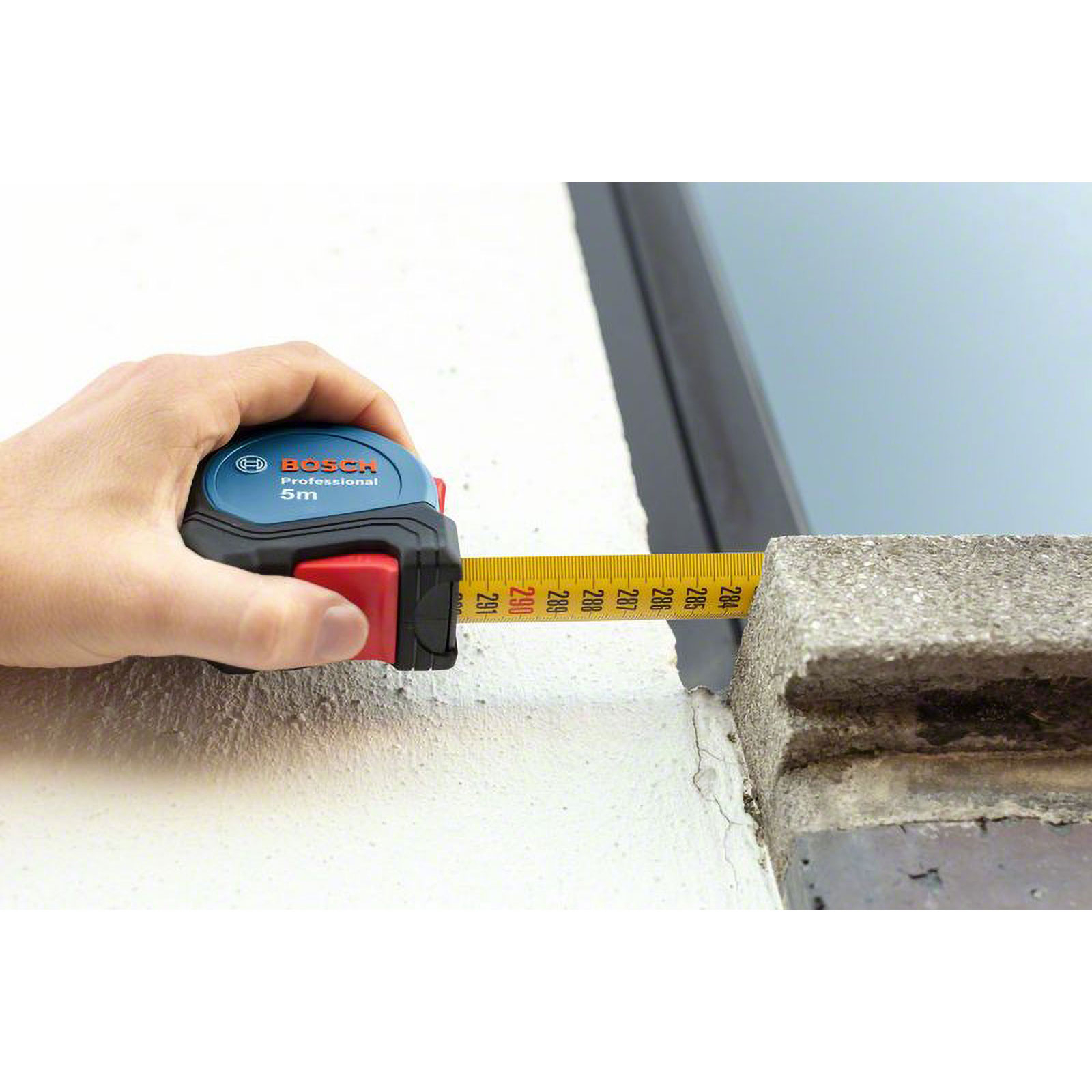El, duvar ve beton blokun üzerinde bir Bosch mezurası tutuyor. Şerit uzatılmış, mesafeyi ölçüyor.