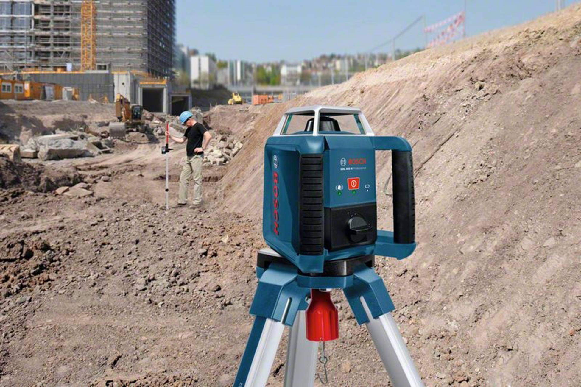 Bir inşaat sahası, tripod üzerinde mavi bir lazer seviye cihazı. Arka planda bir işçi ölçüm çubuğu kullanıyor.