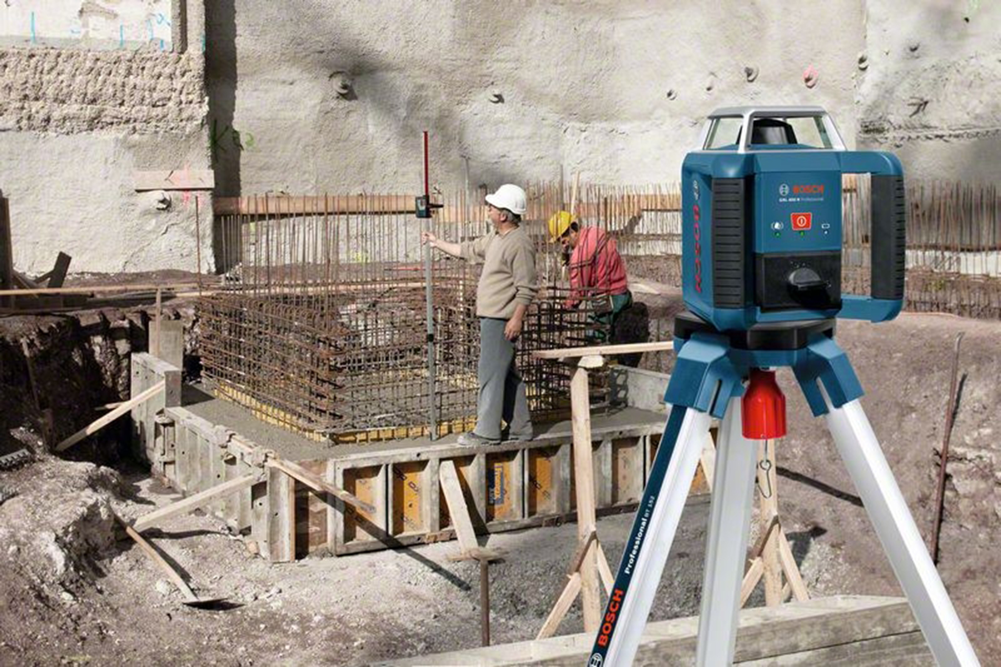 İnşaat işçileri, demir çubuklar ve bir tripod üzerinde mavi bir lazer seviyesi ile inşaat alanı. Arka plan beton bir duvardır.