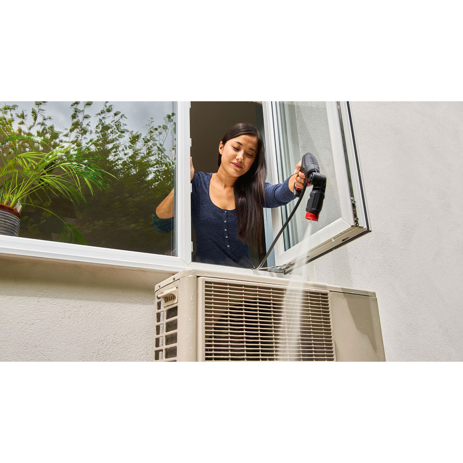 Vrouw maakt airco schoon met hogedrukreiniger. Ze staat bij een raam. Groene planten zijn buiten te zien.