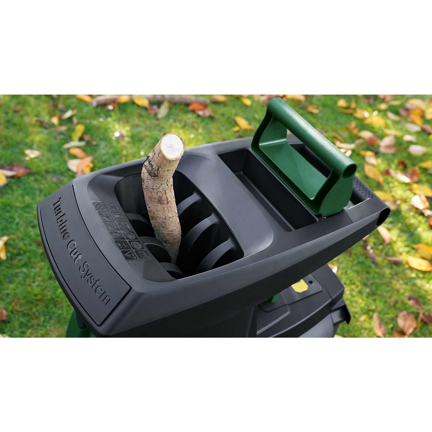 Un biotrituratore nero con manico verde. Viene inserito un tronco di legno. Lo sfondo mostra erba e foglie autunnali.