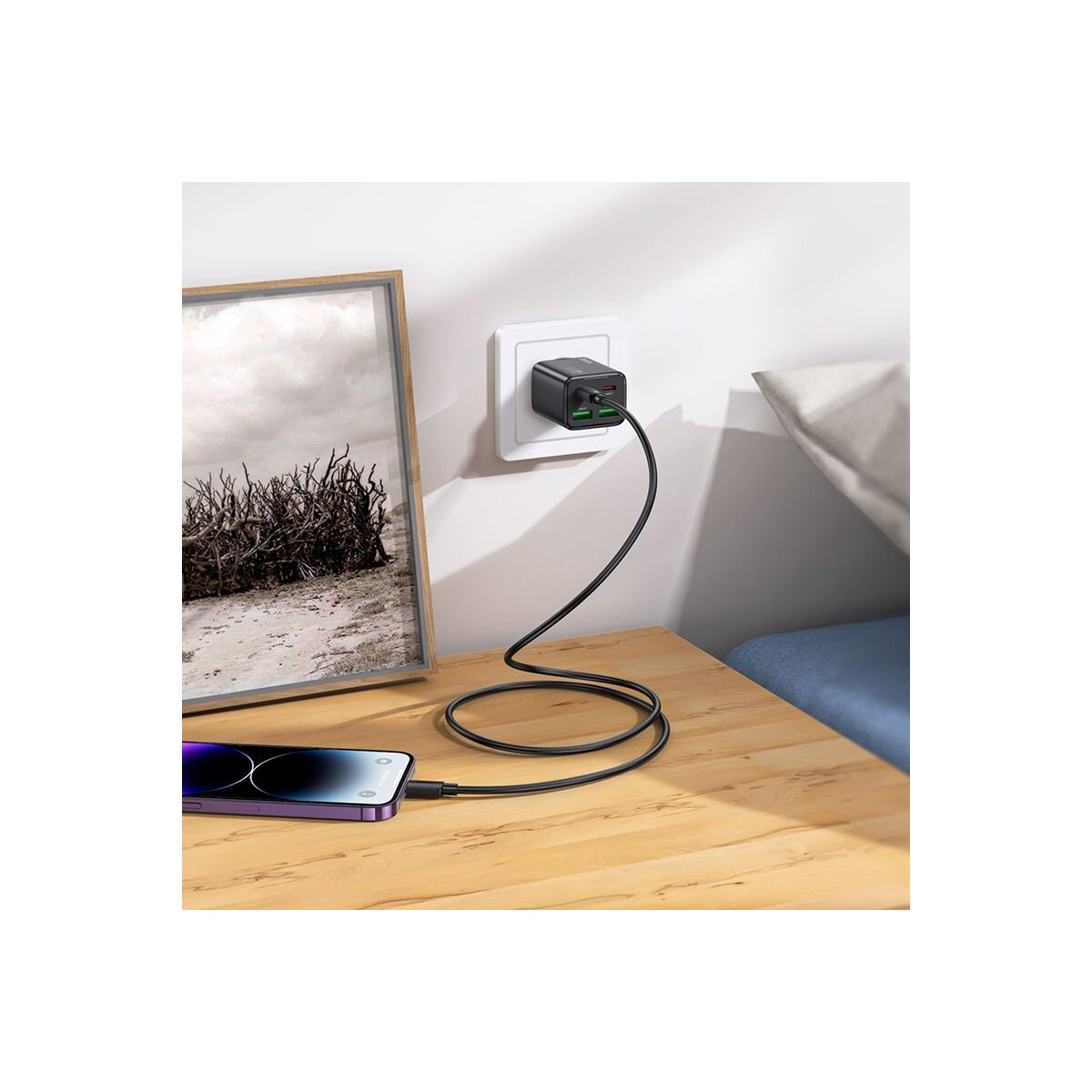 Un téléphone se charge sur une table en bois. Un chargeur est branché dans une prise murale. Une image encadrée est à gauche.