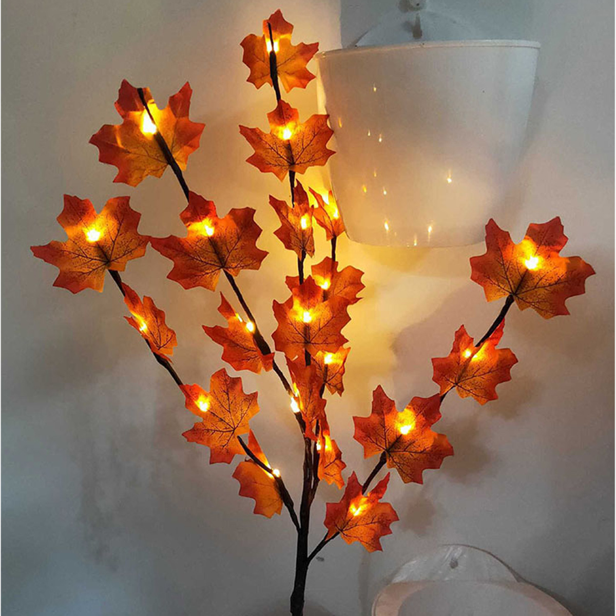 Ein dekorativer Baum mit orangefarbenen Ahornblättern, beleuchtet von kleinen Lichtern. Er steht vor einer weißen Wand mit einem weißen Topf.