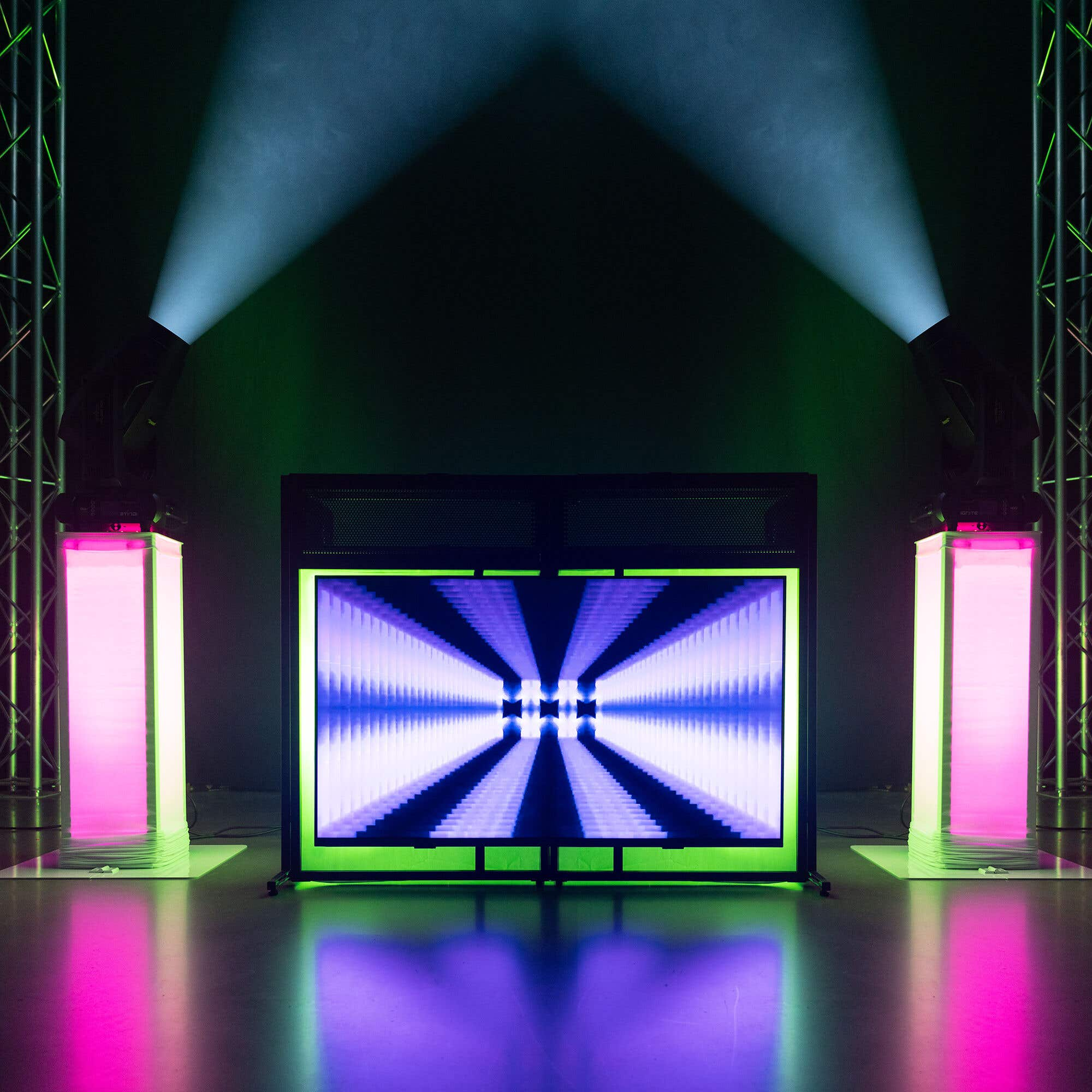 Escenario con pantalla mostrando efecto túnel, flanqueado por pilares de luz y focos. El suelo púrpura refleja luces.
