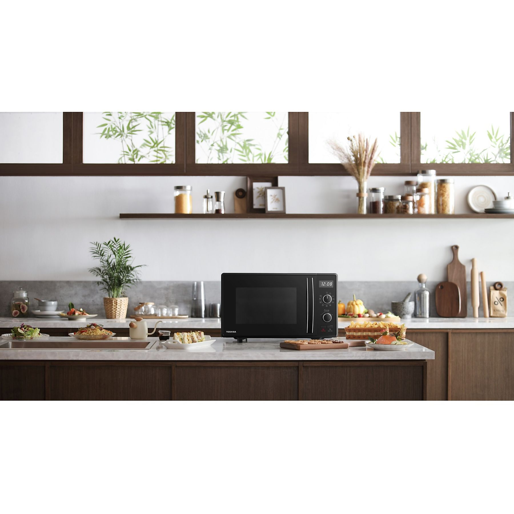 Una scena di cucina con un forno a microonde nero. Cibo e piante sul piano di lavoro e sugli scaffali. Bambù visibile.