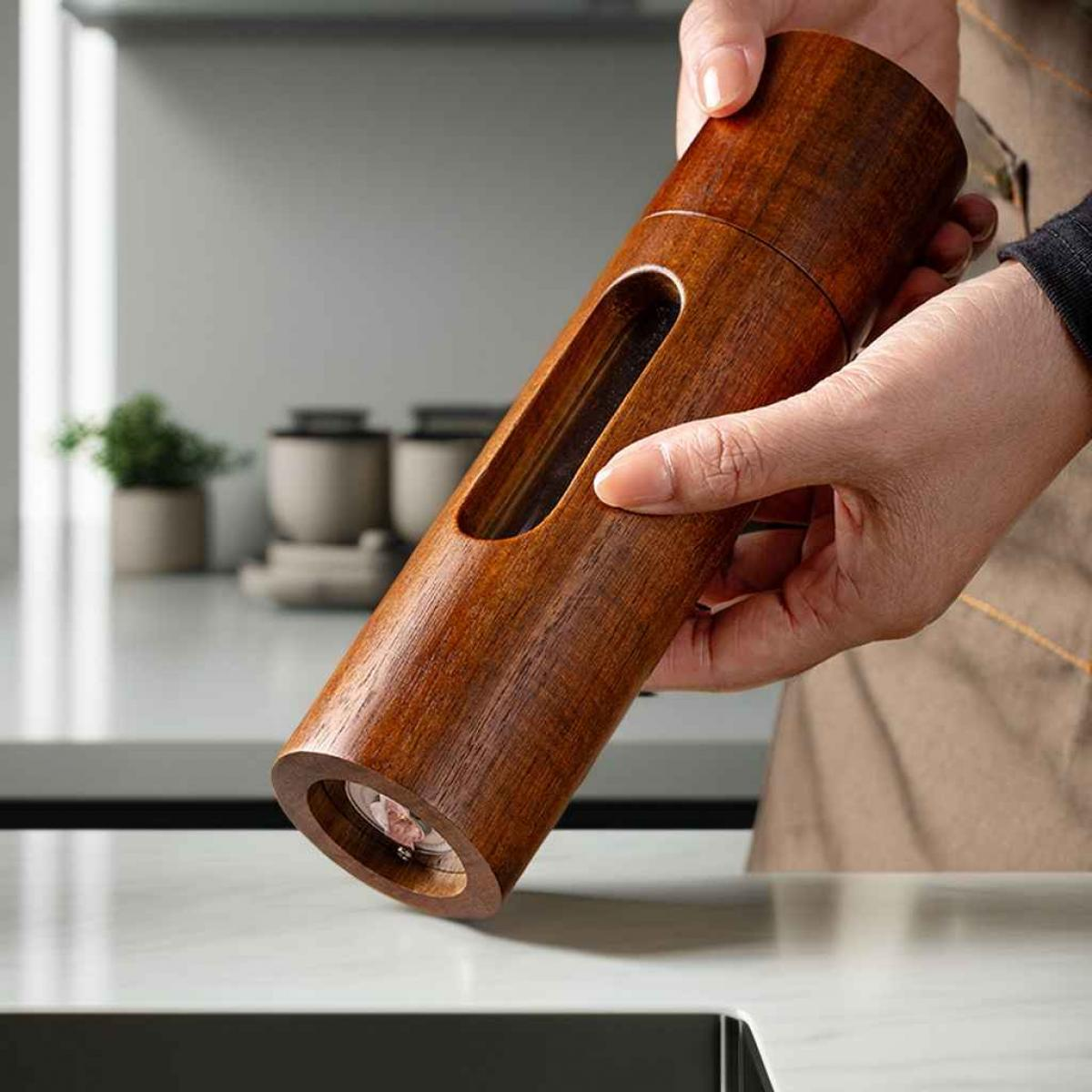 Eine Person hält eine hölzerne Pfeffermühle. Sie hat eine lange, zylindrische Form mit einem Sichtfenster. Der Hintergrund ist eine Küche.