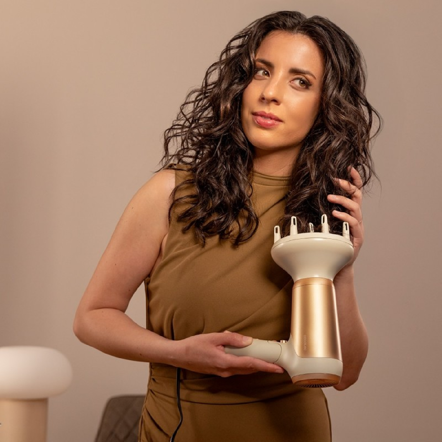 Mujer con cabello rizado sostiene un secador de pelo. Viste un vestido marrón, mirando ligeramente hacia un lado. El fondo es beige.
