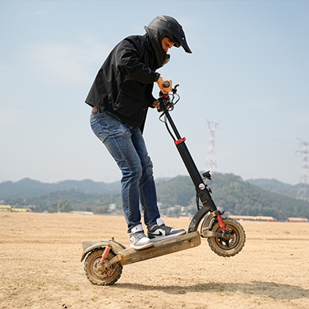 Osoba w kasku jedzie na hulajnodze elektrycznej po polnej ścieżce. Przednie koło hulajnogi jest uniesione. W tle góry i niebo.