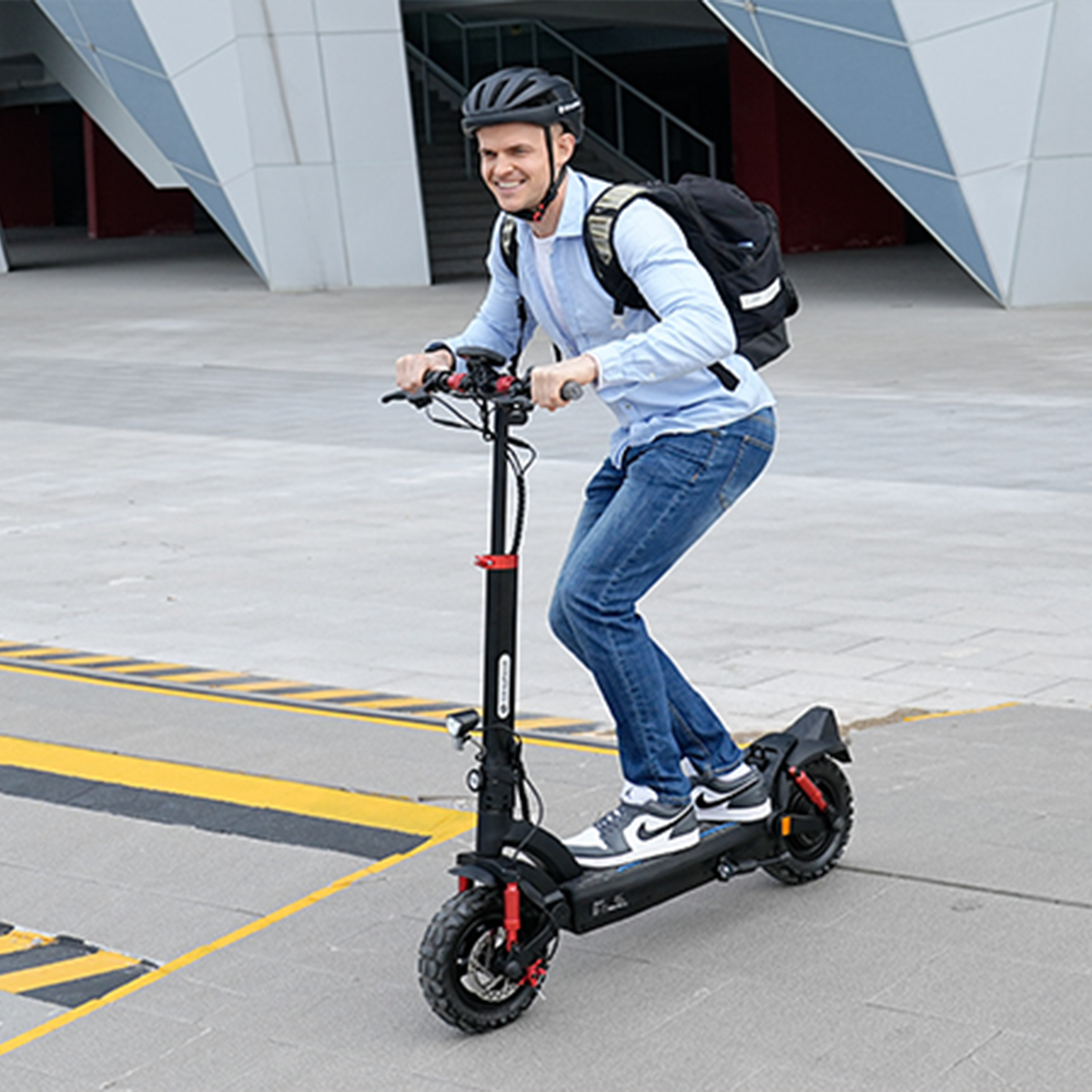 Mężczyzna jedzie na hulajnodze elektrycznej. Ma kask, dżinsy i plecak. Hulajnoga jest czarna z czerwonymi akcentami. Tłem jest budynek.
