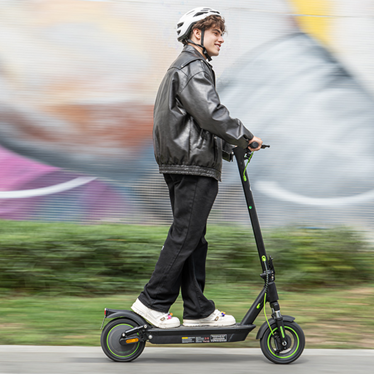Młody mężczyzna jedzie na hulajnodze elektrycznej. Ma na sobie kask, czarną kurtkę i spodnie. Hulajnoga jest czarno-zielona.