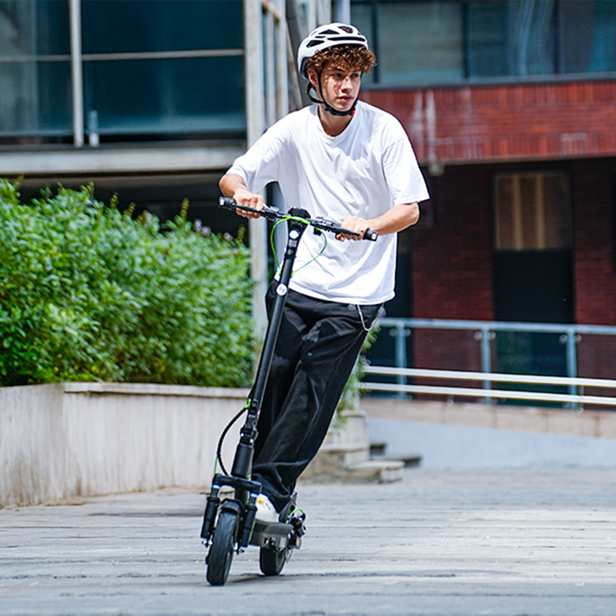 Młody mężczyzna jedzie na hulajnodze elektrycznej. Ma na sobie białą koszulę, czarne spodnie i biały kask. Budynki i zieleń w tle.