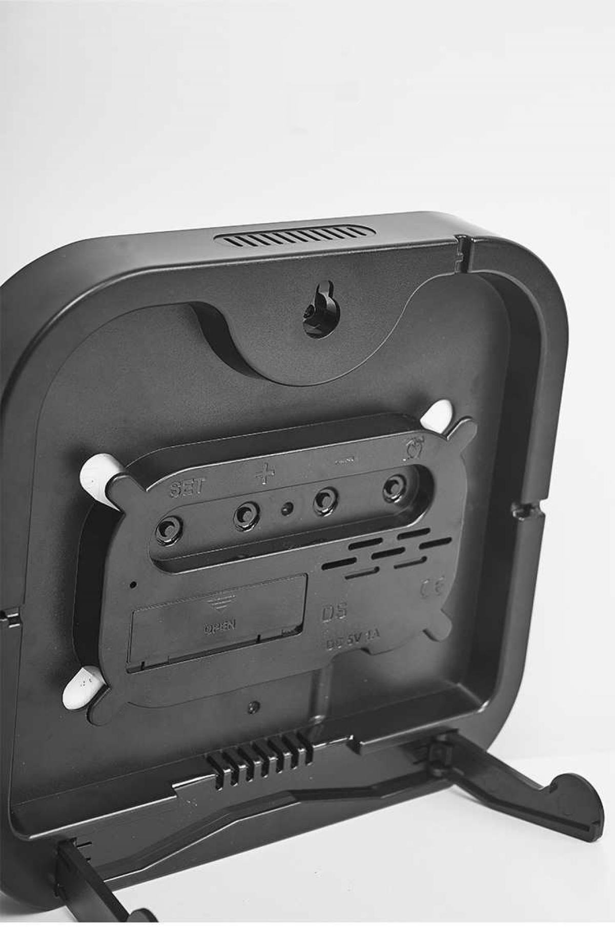 Rückseite einer schwarzen Digitaluhr mit Batteriefach, Knöpfen und Ständer. Details wie Modell DS und DC 5V 1A sind sichtbar.