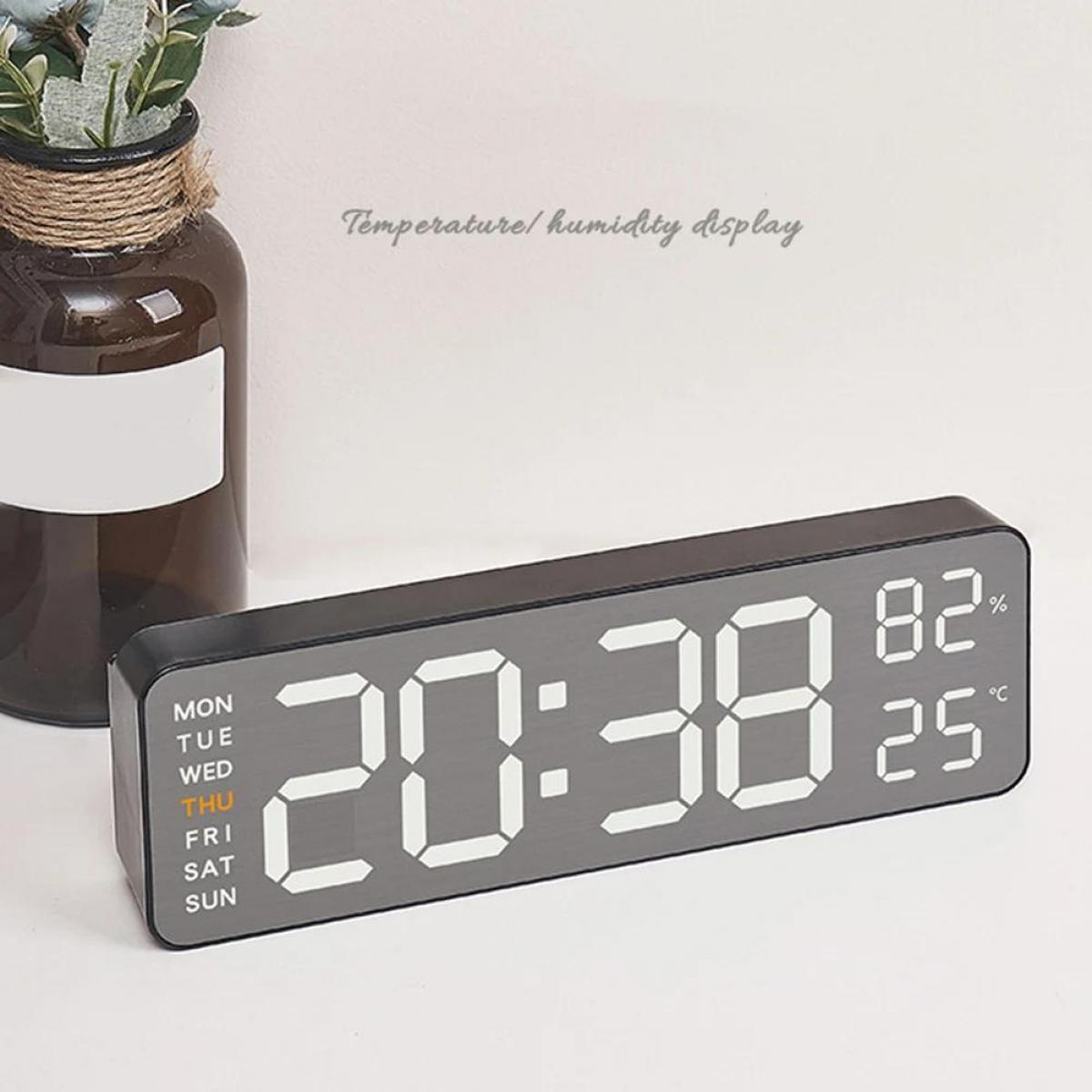 Eine Digitaluhr zeigt 20:38, 82 % Luftfeuchtigkeit und 25 °C an. Links eine Vase mit Pflanzen, der Hintergrund ist weiß.