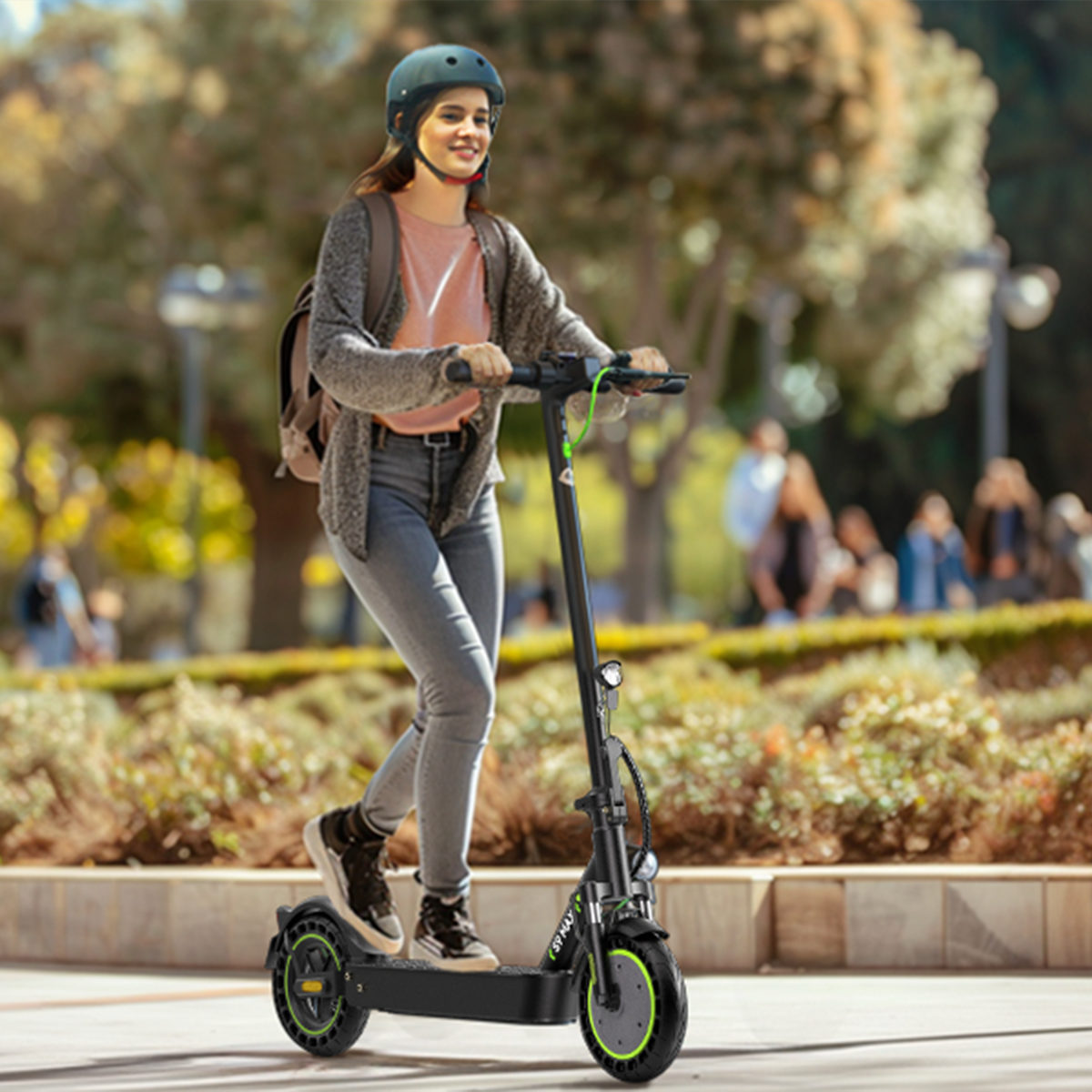 Kobieta w kasku jedzie na hulajnodze elektrycznej po utwardzonej ścieżce. W tle drzewa i ludzie.