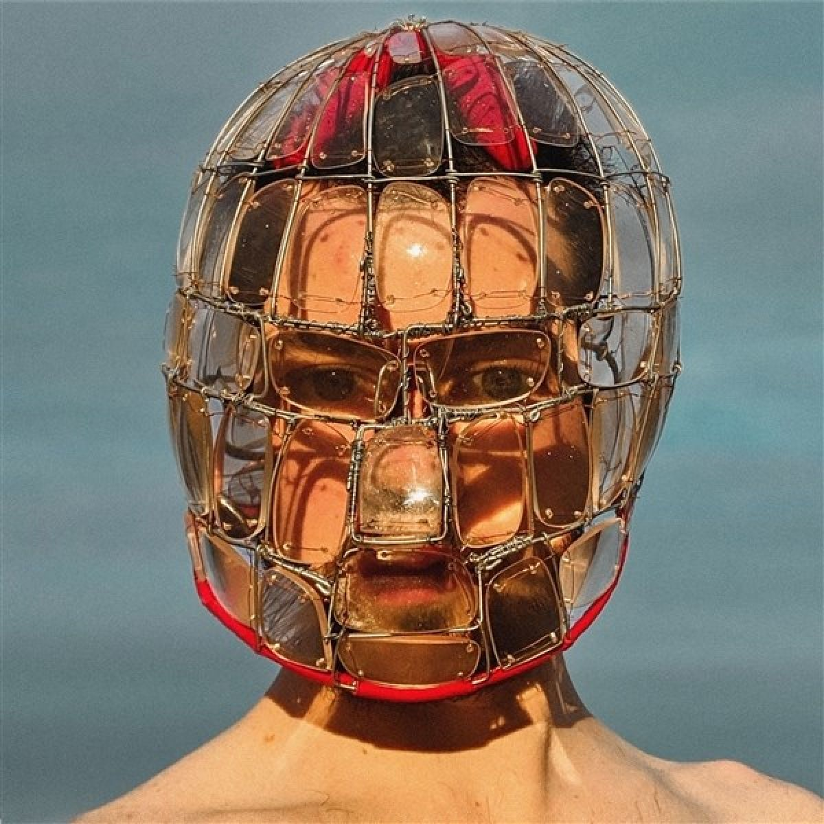 Una persona con il viso coperto da una maschera di filo metallico e vetro. La maschera ha un bordo rosso. Lo sfondo è azzurro.