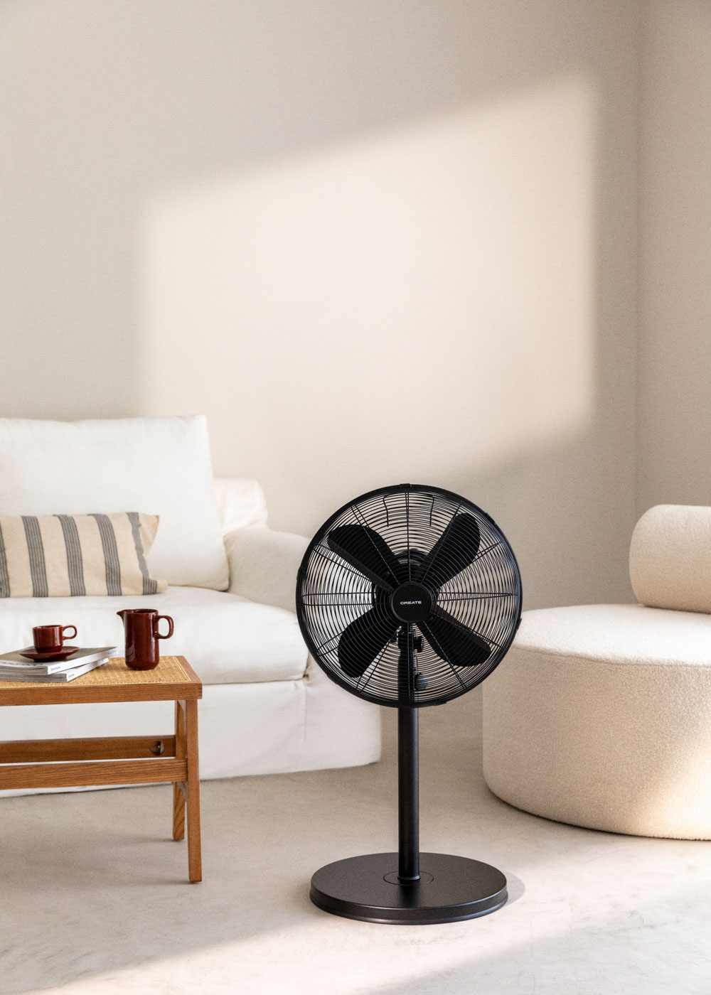 Un ventilador negro está en una sala de estar. Un sofá, una mesita con tazas y una silla son visibles.