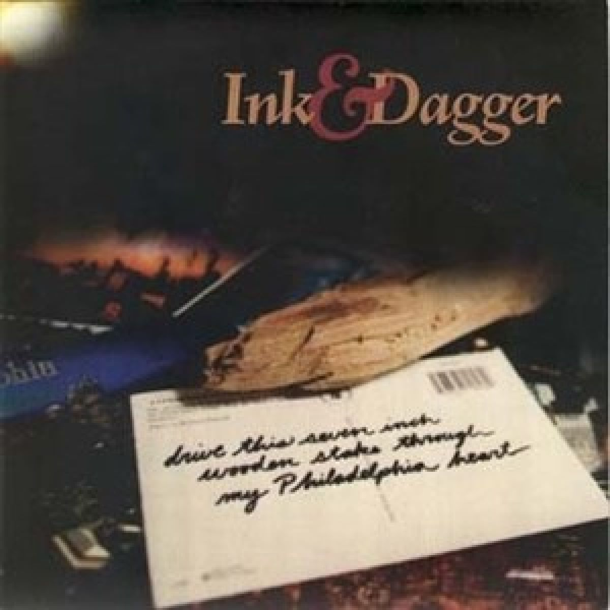 Albumhoes met tekst 'Ink & Dagger'. Een houten voorwerp en een ansichtkaart met handgeschreven tekst op de voorgrond.