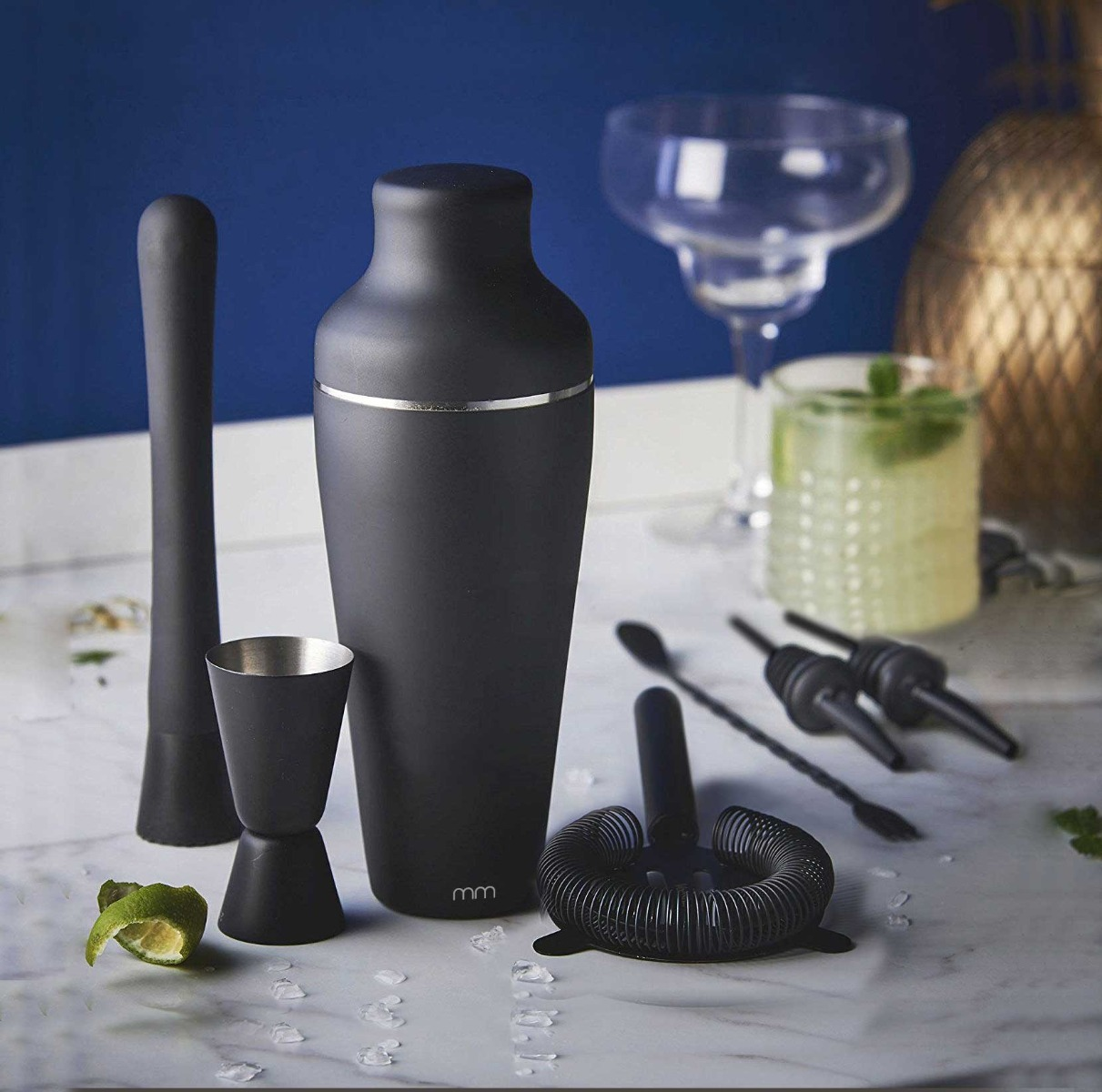 A cocktail set on a marble surface, with a shaker, jigger, muddler, strainer, spoons, and a glass with a cocktail.