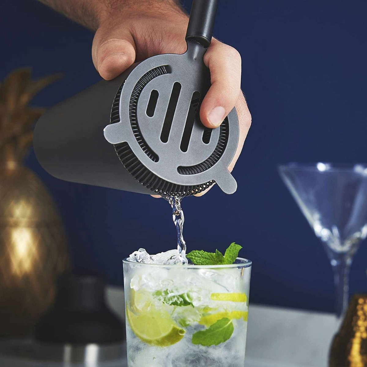 A hand pouring liquid from a gray cocktail strainer into a glass with ice, lime, and mint. Dark blue background.