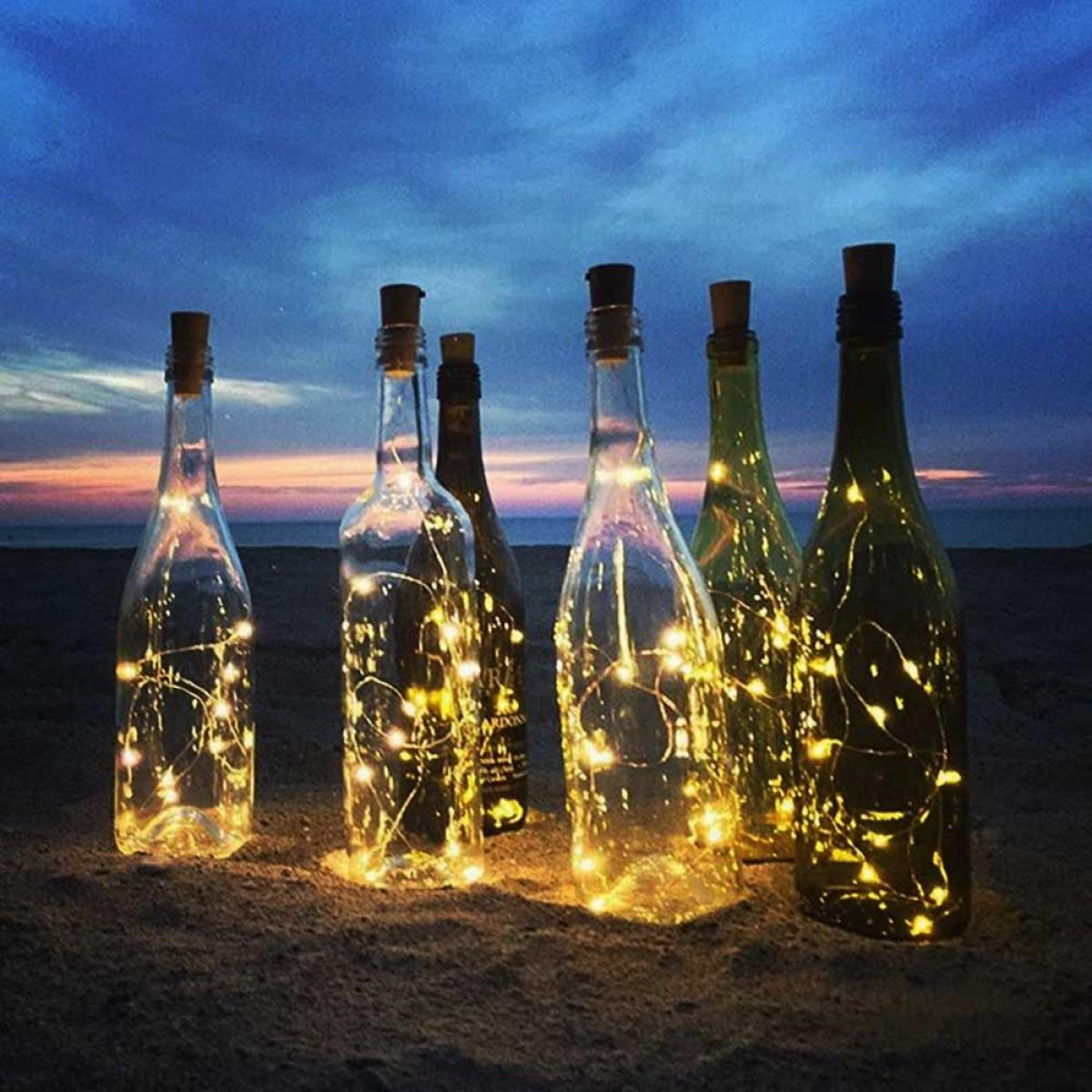 Six lit bottles on sand at dusk. Clear and green bottles with fairy lights and corks. Sunset in the background.