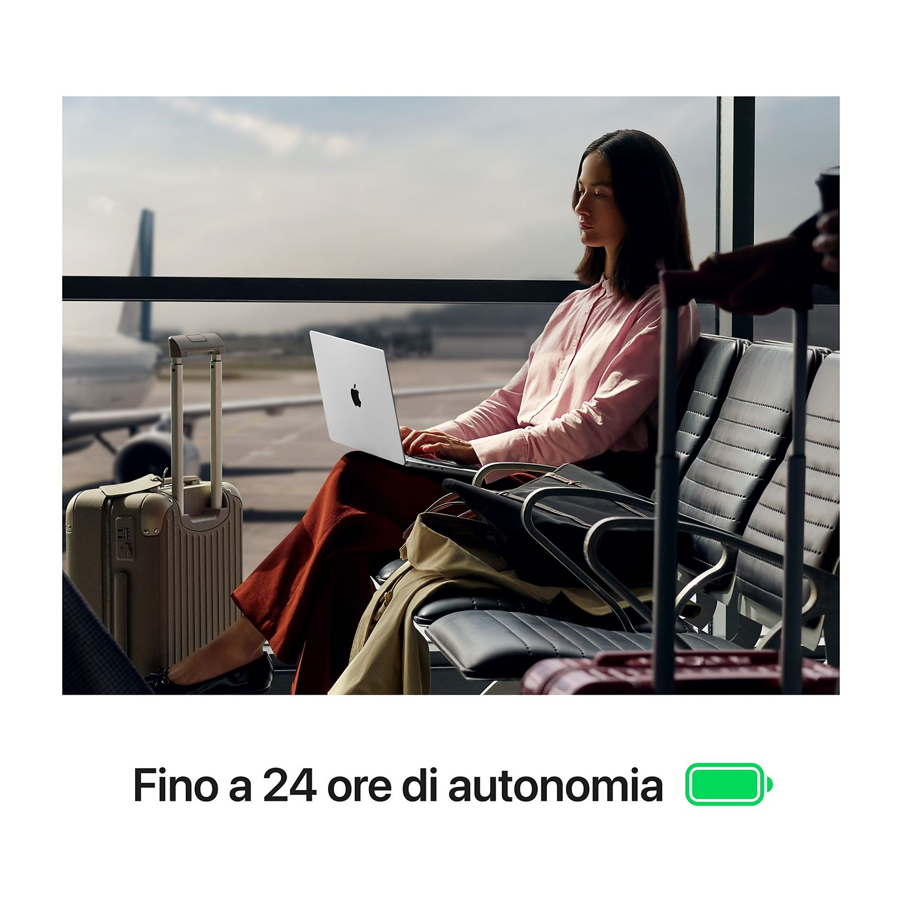 Donna con laptop in aeroporto. Bagagli vicini. Aereo visibile dalla finestra. Icona batteria in basso.