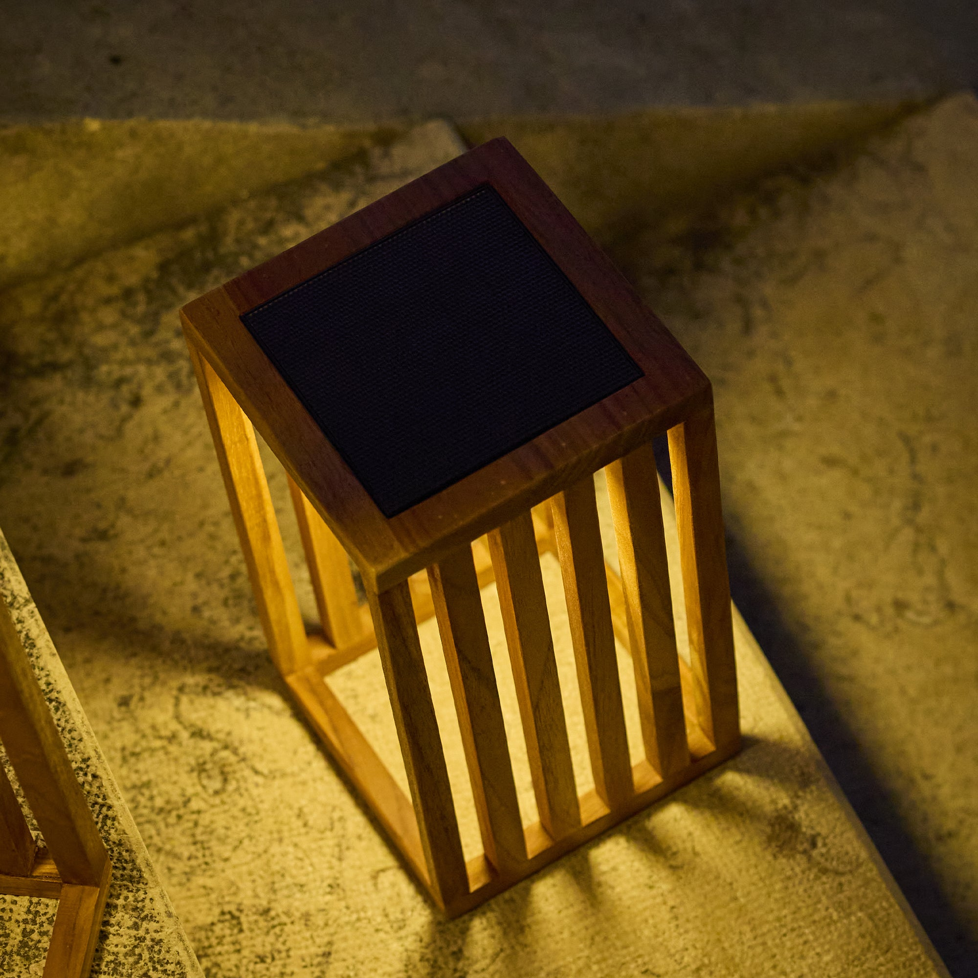 Eine Holzlampe mit schwarzer Oberseite steht auf einer strukturierten Oberfläche. Das Licht scheint durch die Lamellen.