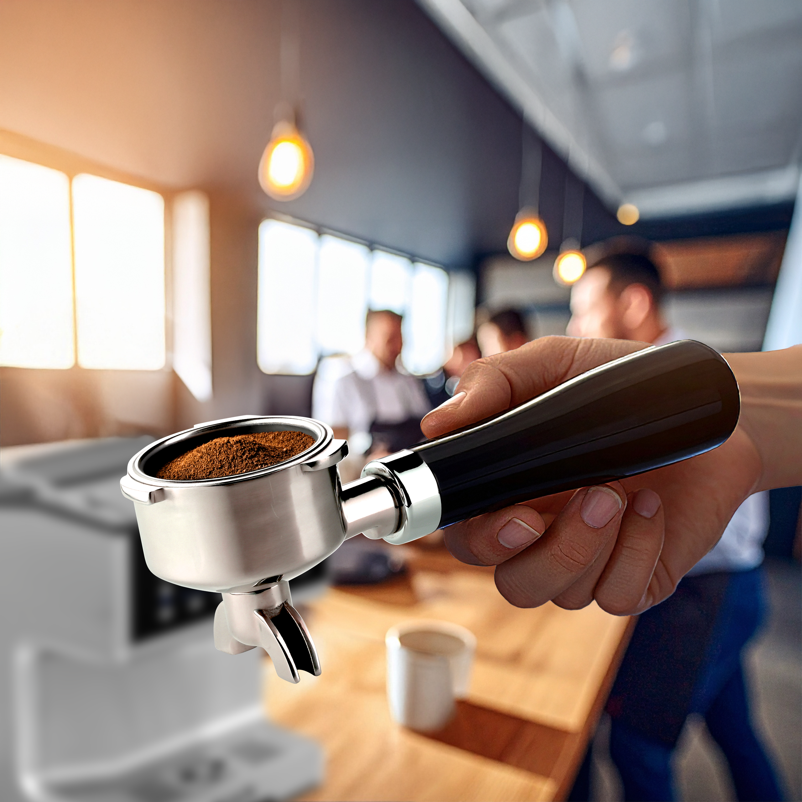 Ręka trzyma kolbę z mieloną kawą. W tle rozmyta scena kawiarni.