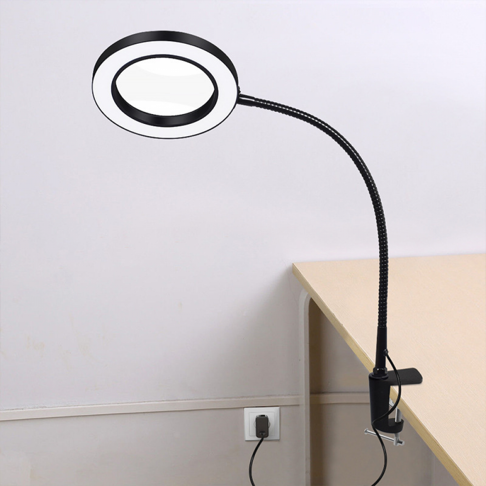 Eine Lupe mit kreisförmiger Beleuchtung wird an einem Schreibtisch befestigt. Im Hintergrund ist eine Wand mit Steckdose zu sehen.