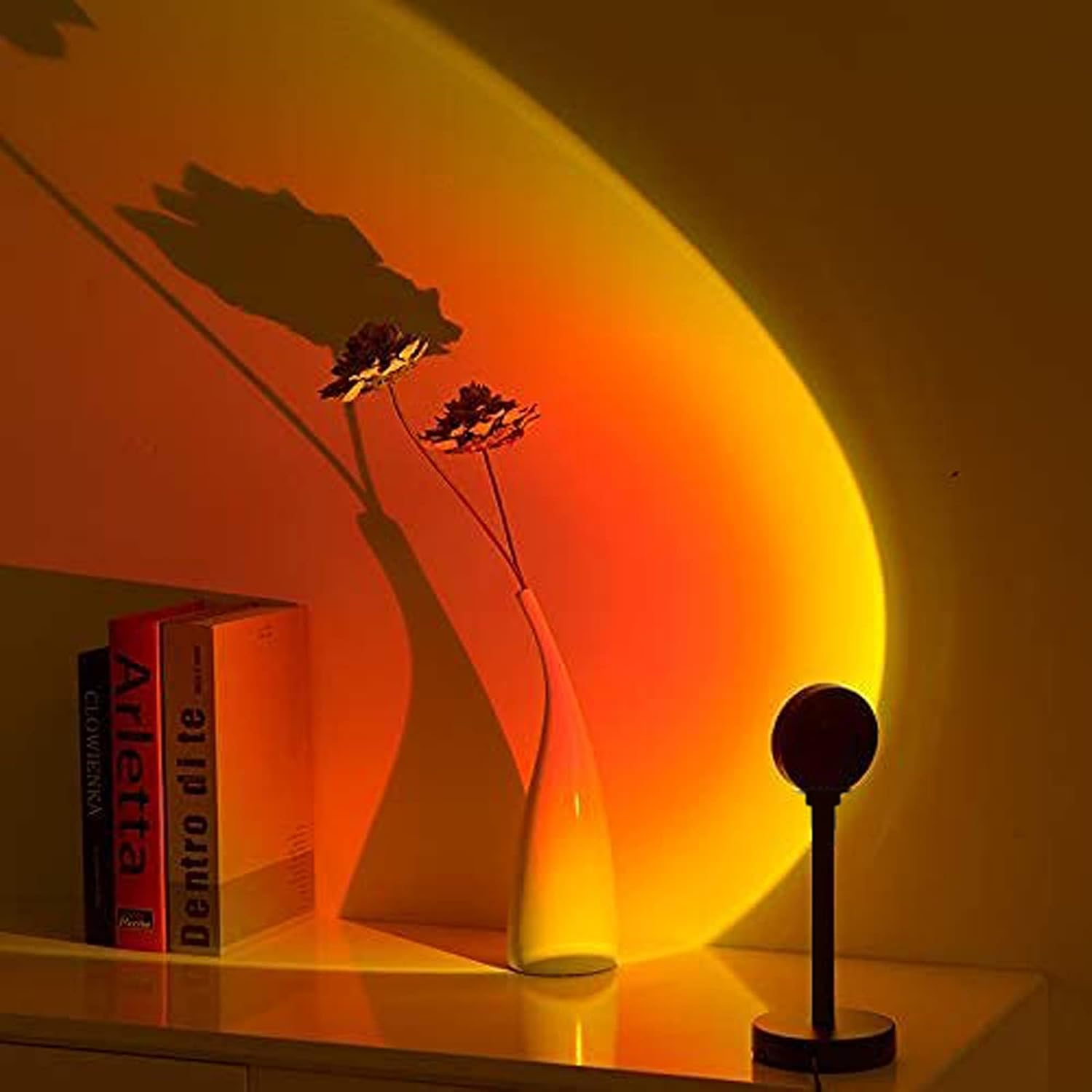 Eine Lampe projiziert einen gelb-orangen Kreis an die Wand. Eine Vase mit Blumen und Bücher stehen auf einem Tisch. Die Szene ist beleuchtet.