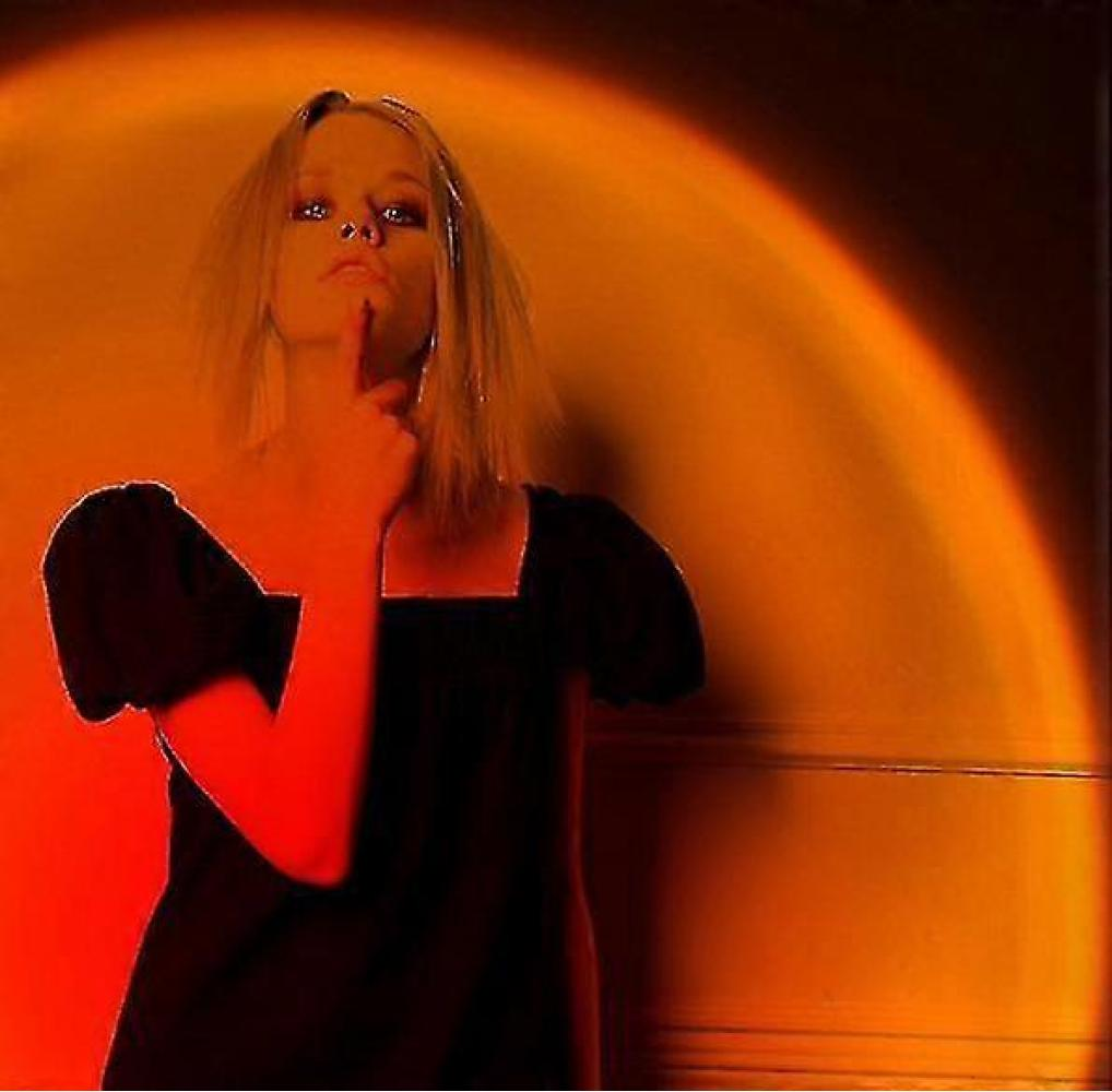 Blonde Frau in schwarzem Kleid, Finger an der Lippe. Oranges Licht im Hintergrund. Dunkle Wand.